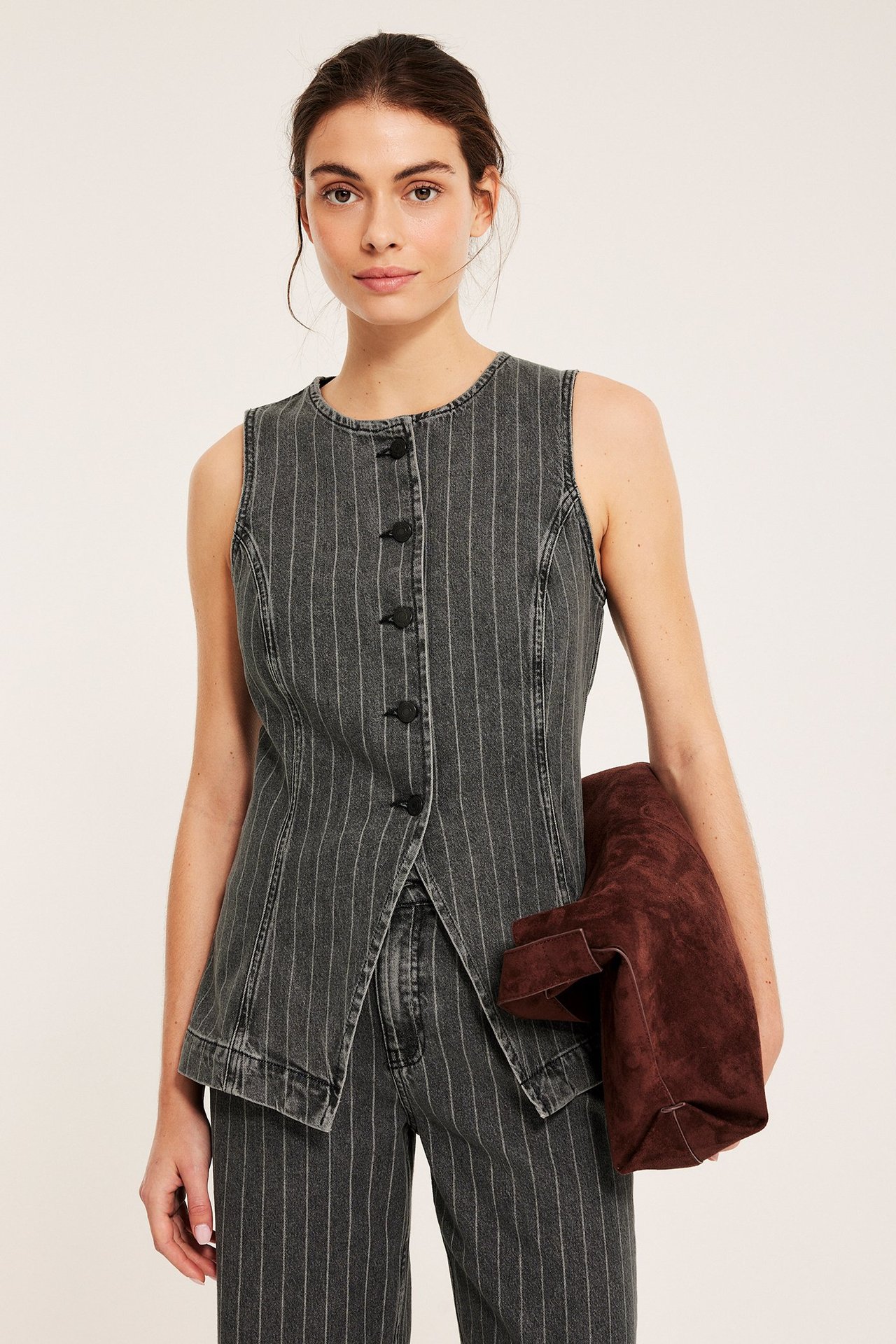 Forfra: Kvinne i figursydd mørkegrå stripet denimvest, matchende jeans, holder brun veske.
