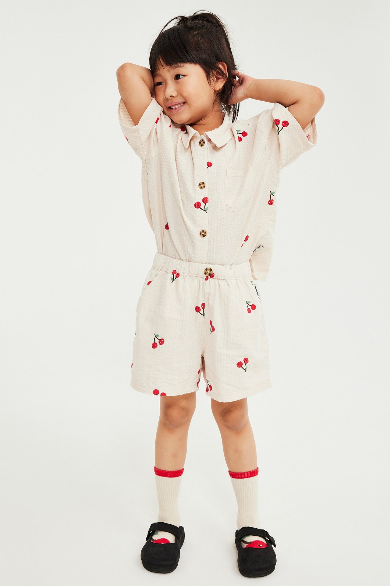 Widok z przodu: Dziecko w beżowej koszuli z bawełny fakturowanej w wiśnie i pasujących szortach, białych skarpetkach, czarnych butach.