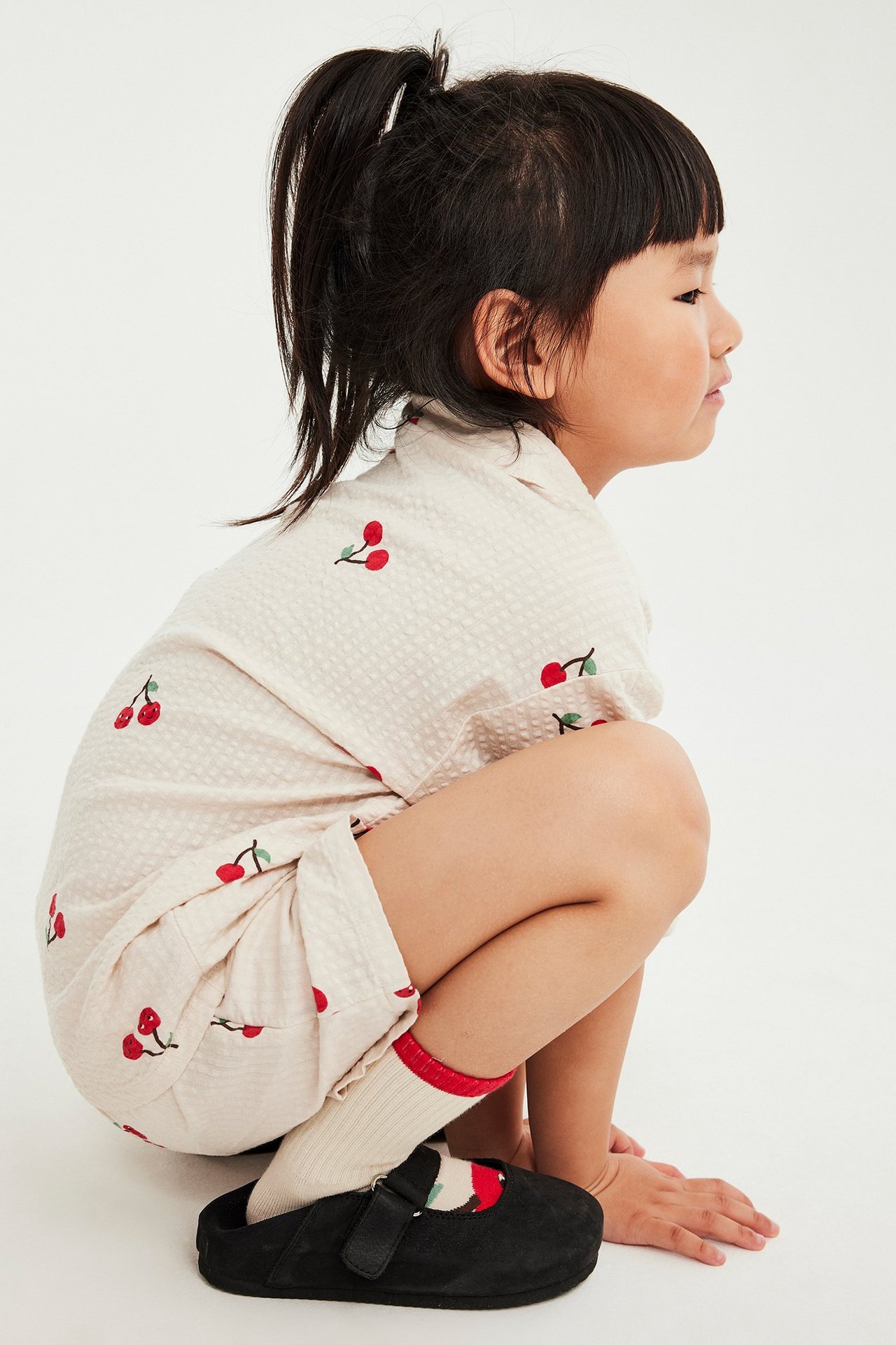 Widok z boku: dziecko w beżowym zestawie seersucker w wiśnie, z beżowymi skarpetkami i czarnymi butami.