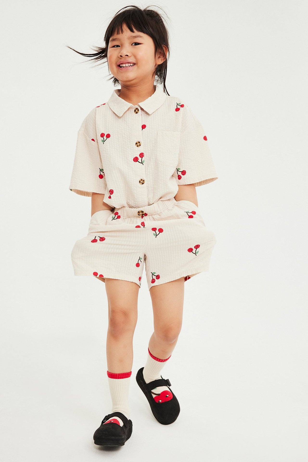 Framifrån: Kid i beige körsbärsmönstrad skjorta med knappar och matchande shorts i seersucker, plus gräddvita strumpor och svarta skor.