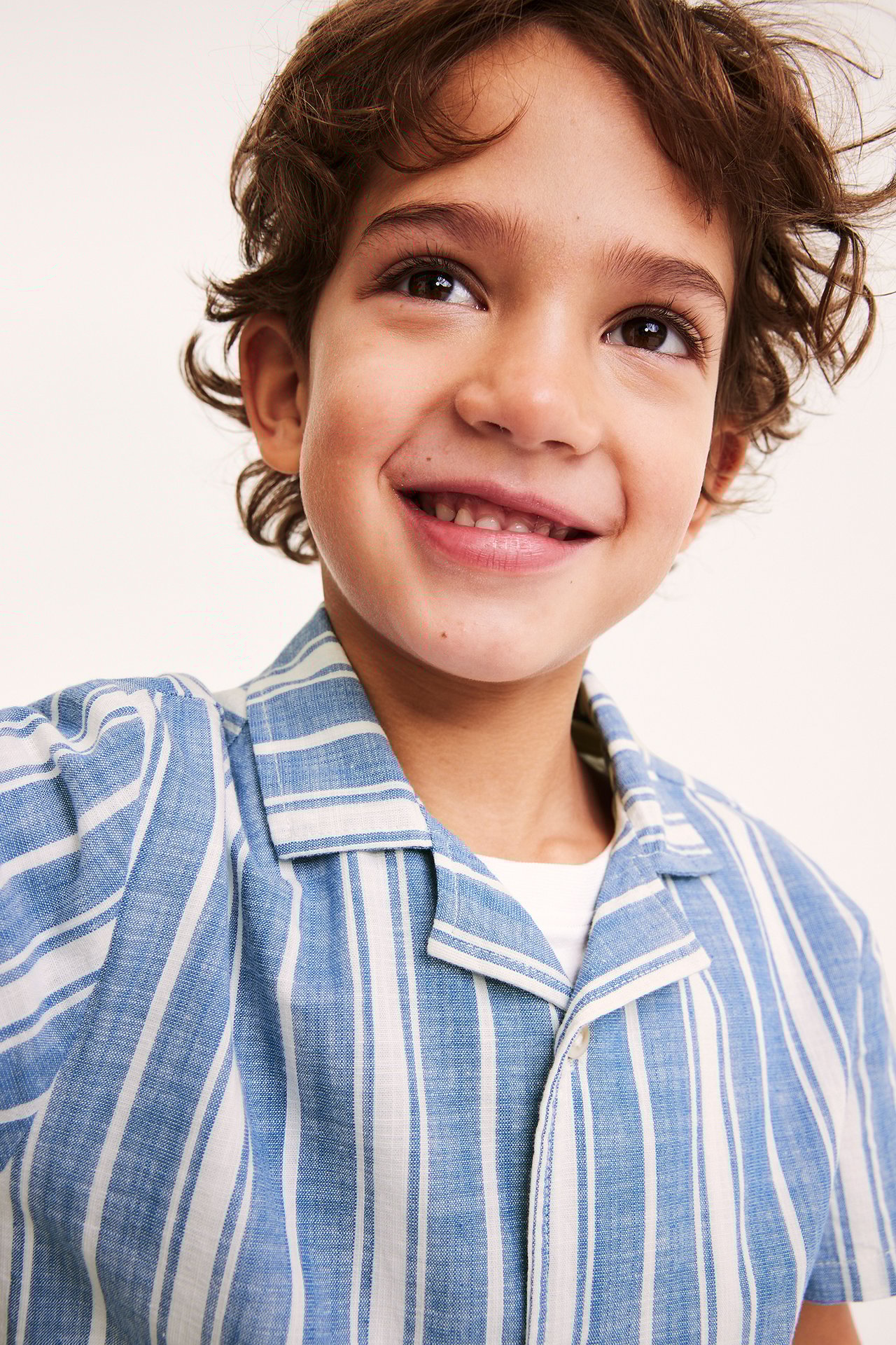 Edestäpäin, hymyilevä lapsi yllään raidallinen sininen lyhythihainen puuvillapaita valkoisen t-paidan päällä.