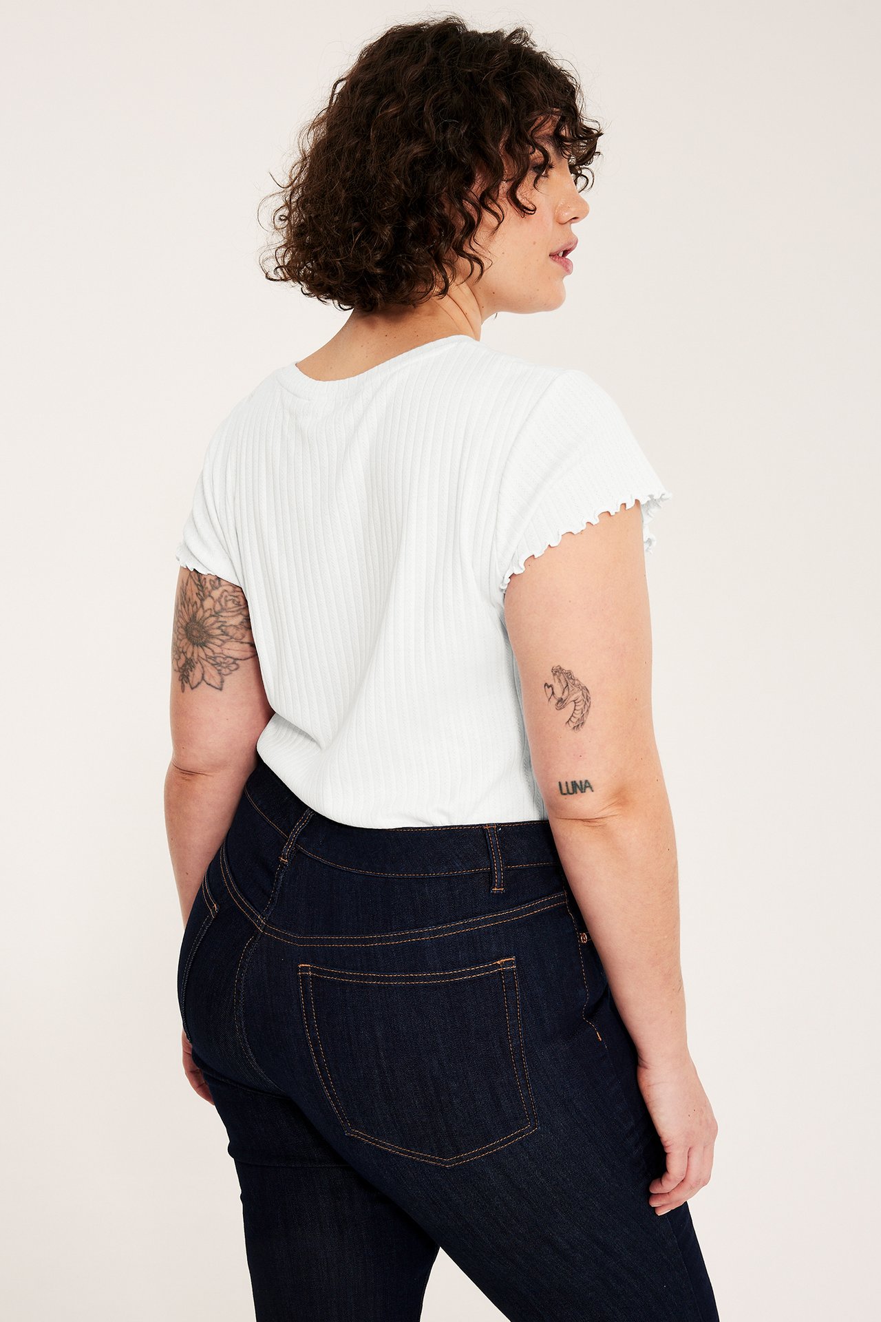 Kobieta od boku w białym, dopasowanym, strukturalnym bawełnianym t-shircie z falistymi rękawami i ciemnoniebieskich dżinsach.