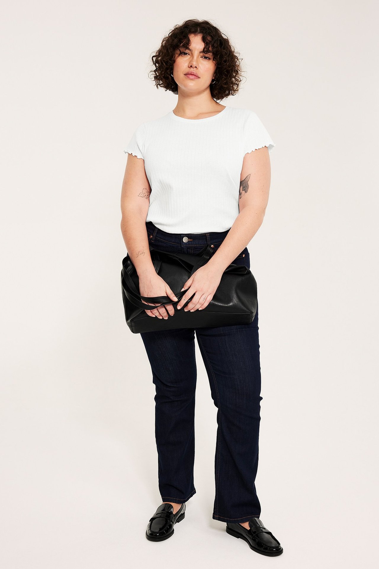 En kvinne har på seg en hvit pointelle baby-t-skjorte, mørkeblå jeans, svarte loafers og holder en svart veske.