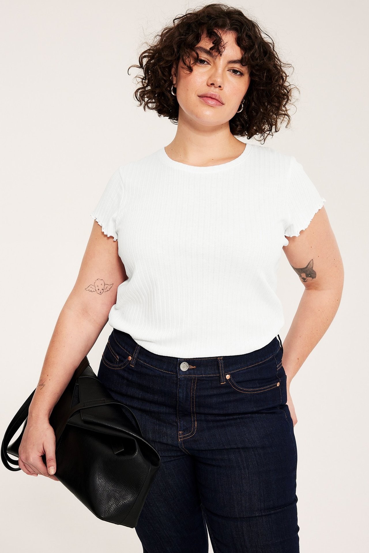 Frontutsikt: Kvinne i hvit ribbet baby-t-skjorte med babylock-ermer, mørkeblå jeans, med svart håndveske.