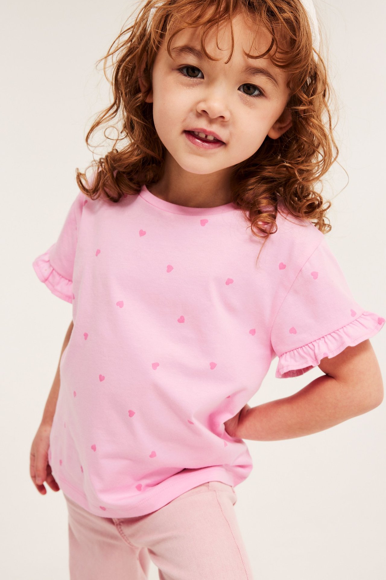 Framifrån: Ett barn bär en ljusrosa bomulls-t-shirt med hjärtmönster, volangärmar och rosa byxor.