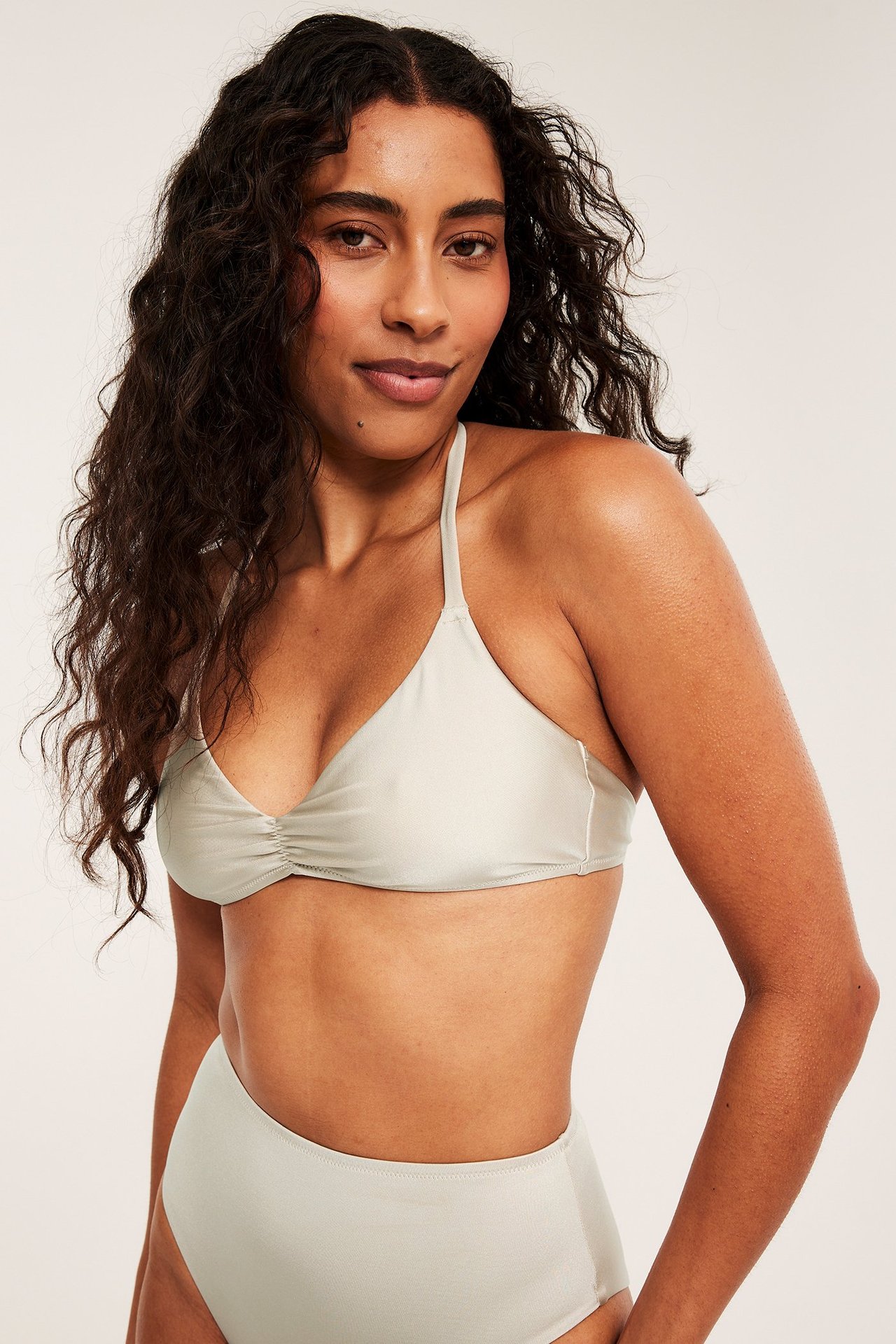 Naisen etukuva beigessä topatussa kolmioliivi-bikiniyläosassa ja yhteensopivassa korkeavyötäröisessä alaosassa.