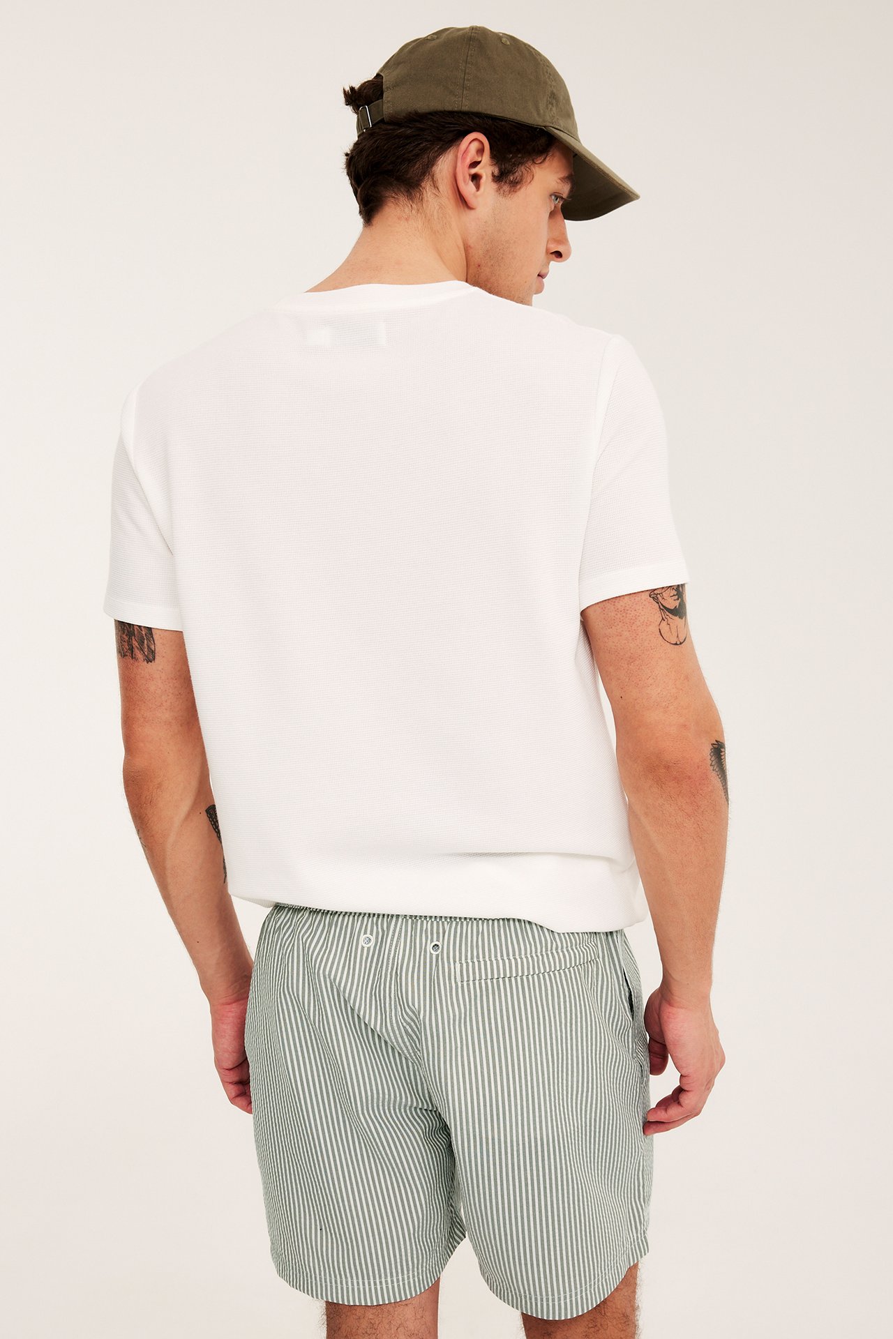 Widok z tyłu mężczyzny w ciemnozielonych, pasiastych szortach z seersuckera, białym T-shircie i zielonej czapce.