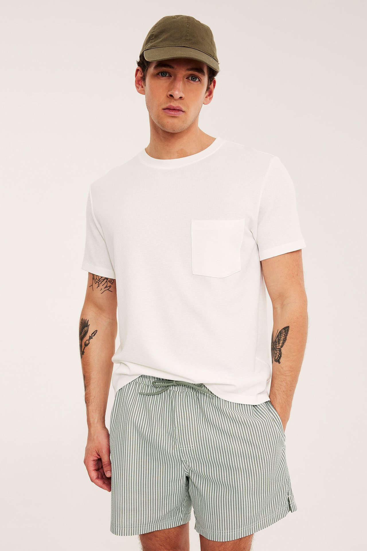 Frontvy av man i grönrandiga bäckebölja-badshorts, vit t-shirt och olivgrön keps.