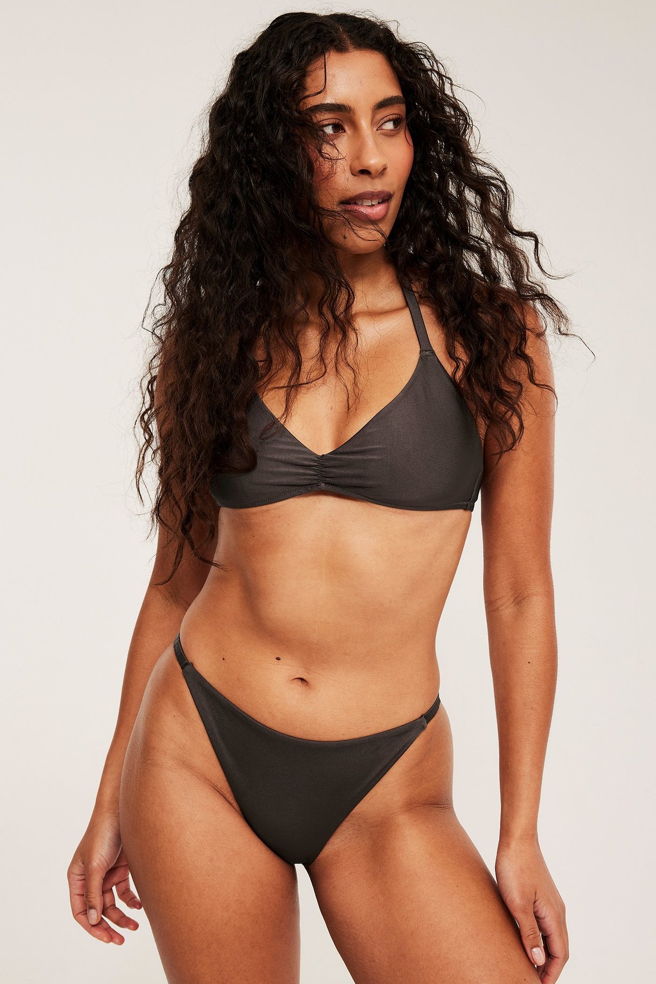 Edestäpäin nainen tummanruskeissa brazilian-bikinihousuissa ja rypytetyssä halterneck-bikinitopissa.