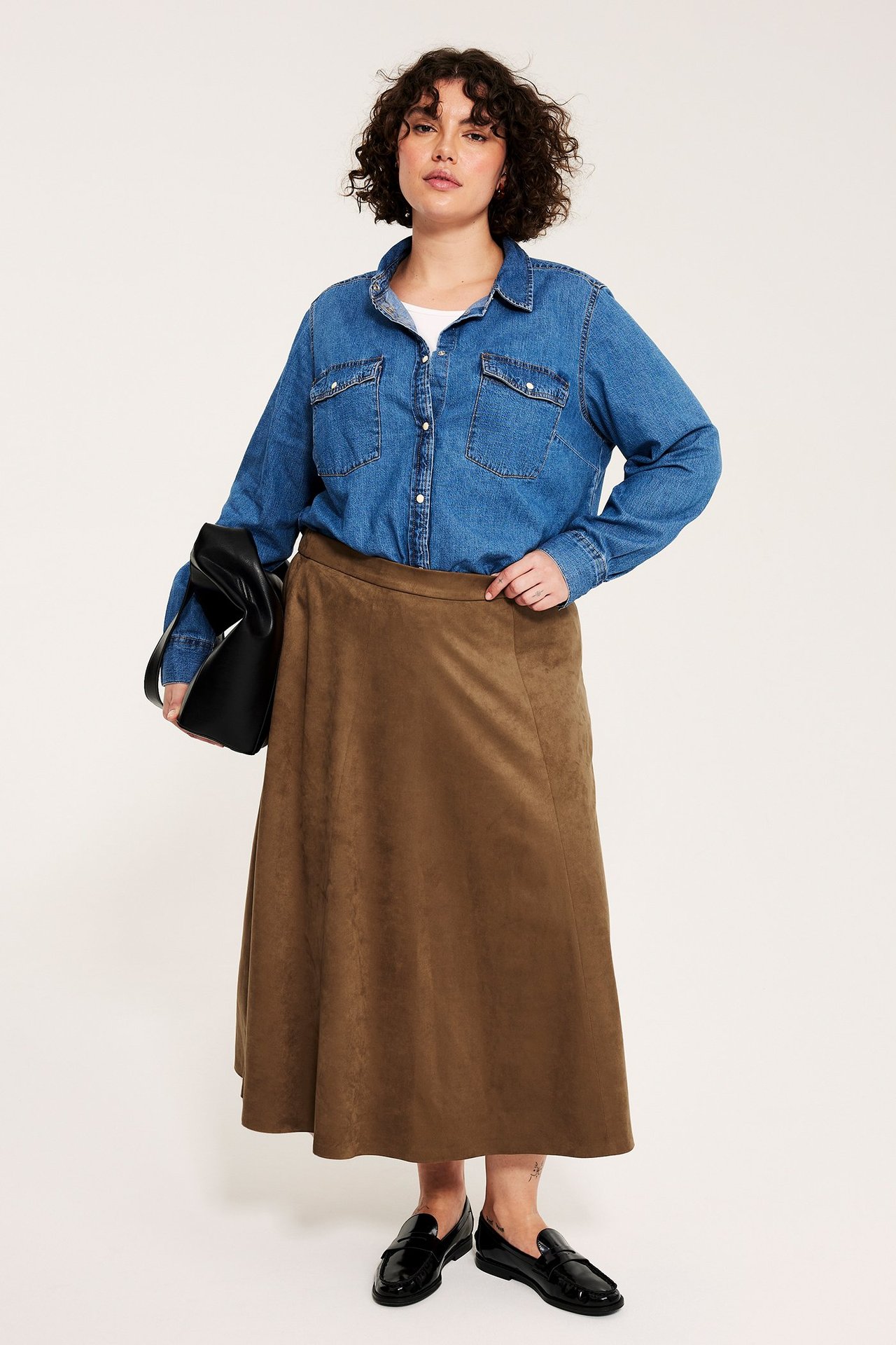 Edestä: nainen pitkässä ruskeassa tekomokkahameessa, farkkupaidassa, mustat loaferit, pitelee mustaa laukkua.