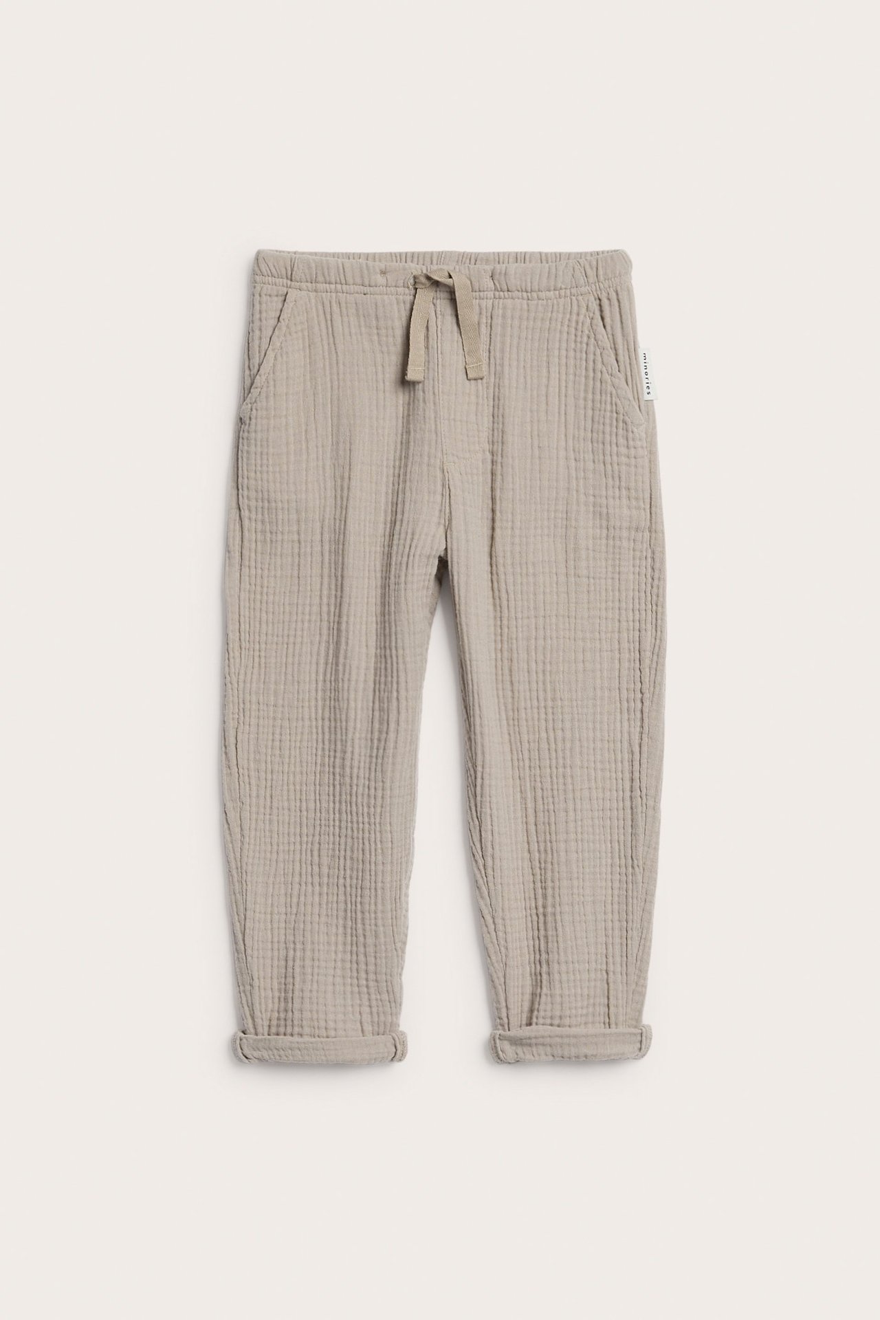 Widok z przodu: beżowe, jednolite spodnie muślinowe dla dziecka z kieszeniami, imitacją sznurka i podwiniętymi nogawkami.