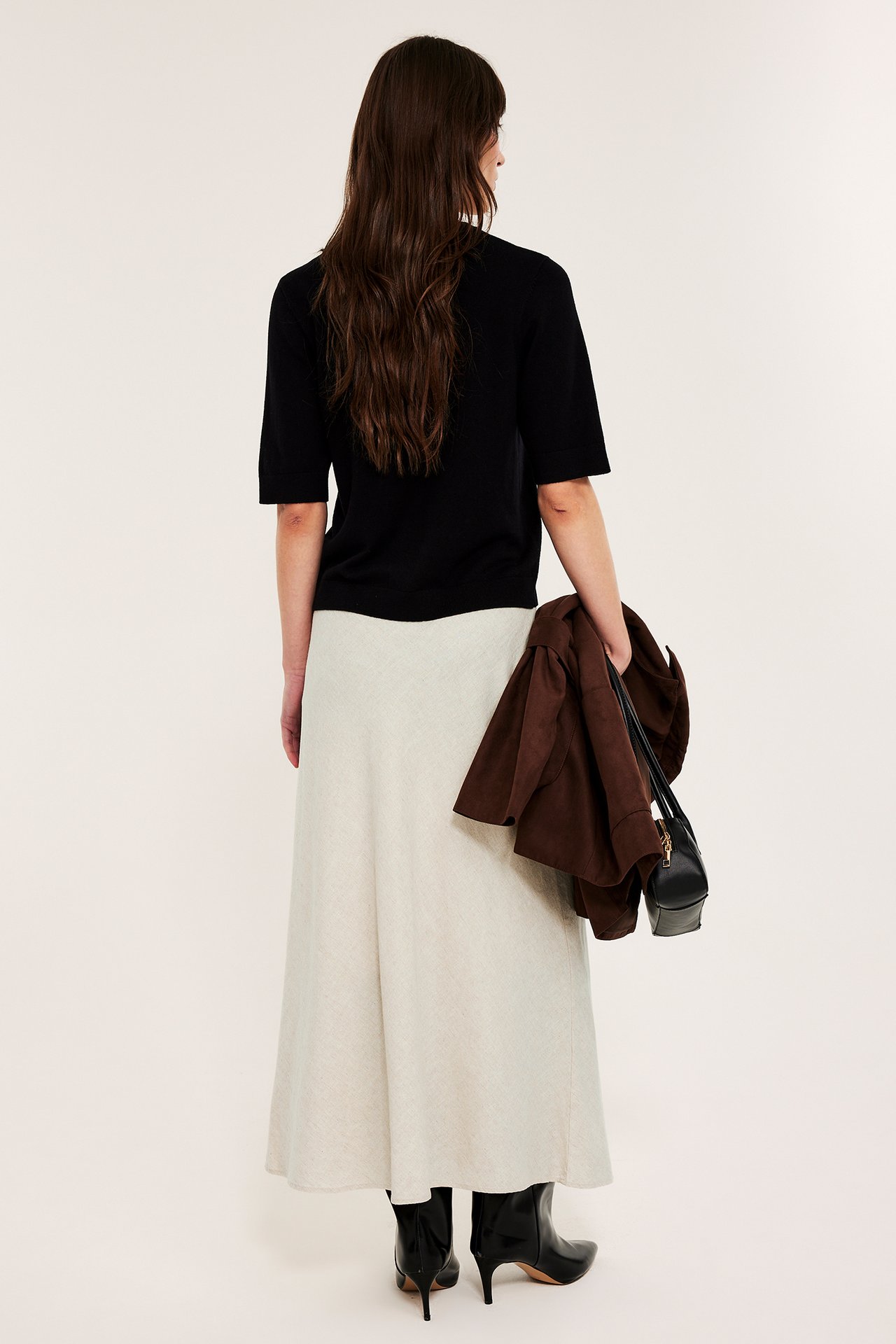 Rygg: Kvinne i beige lin-midiskjørt, svart topp, svarte støvler, holder brun jakke og svart veske.