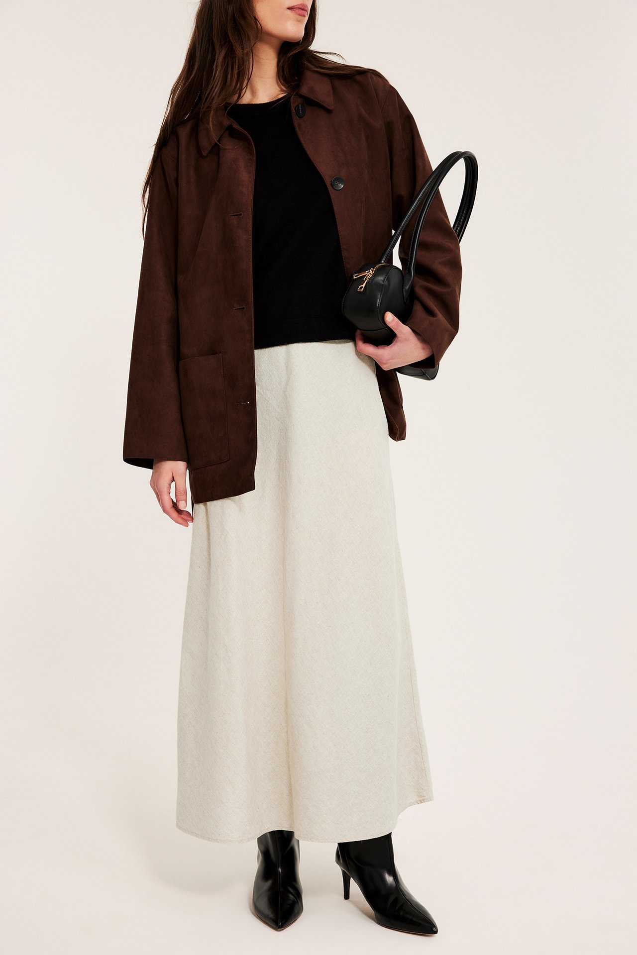 Frontalvisning: kvinne i beige lin-midiskjørt, svart topp, brun jakke, svarte støvler, svart veske.