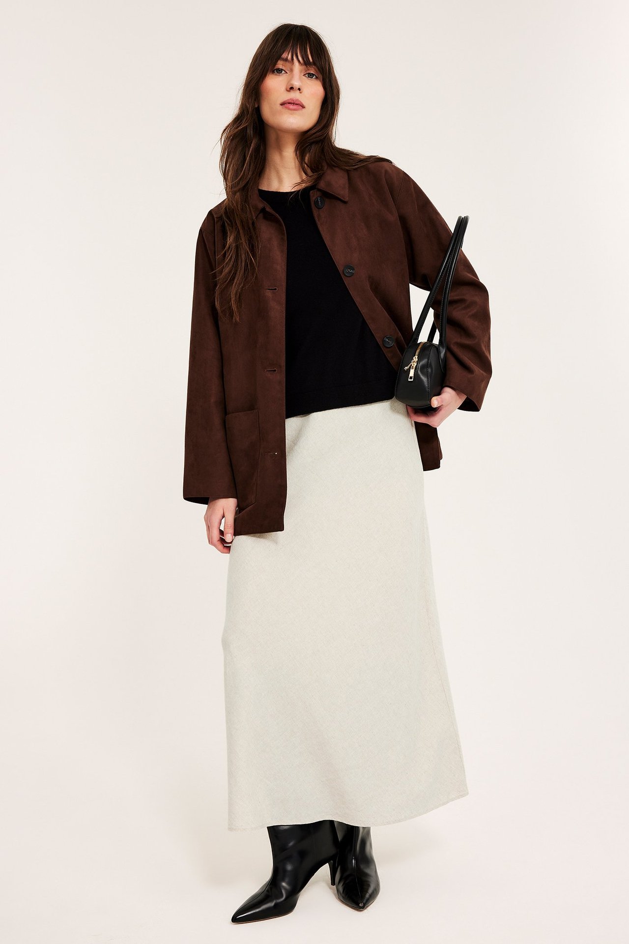 Kobieta w beżowej spódnicy midi z lnu, brązowej kurtce, czarnym topie, czarnych kozakach, trzyma czarną torebkę.