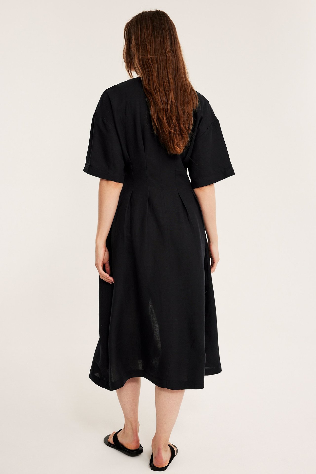 Naisen takakuva mustassa pellavasekoitemekossa, jossa leveät hihat ja mustat sandaalit.