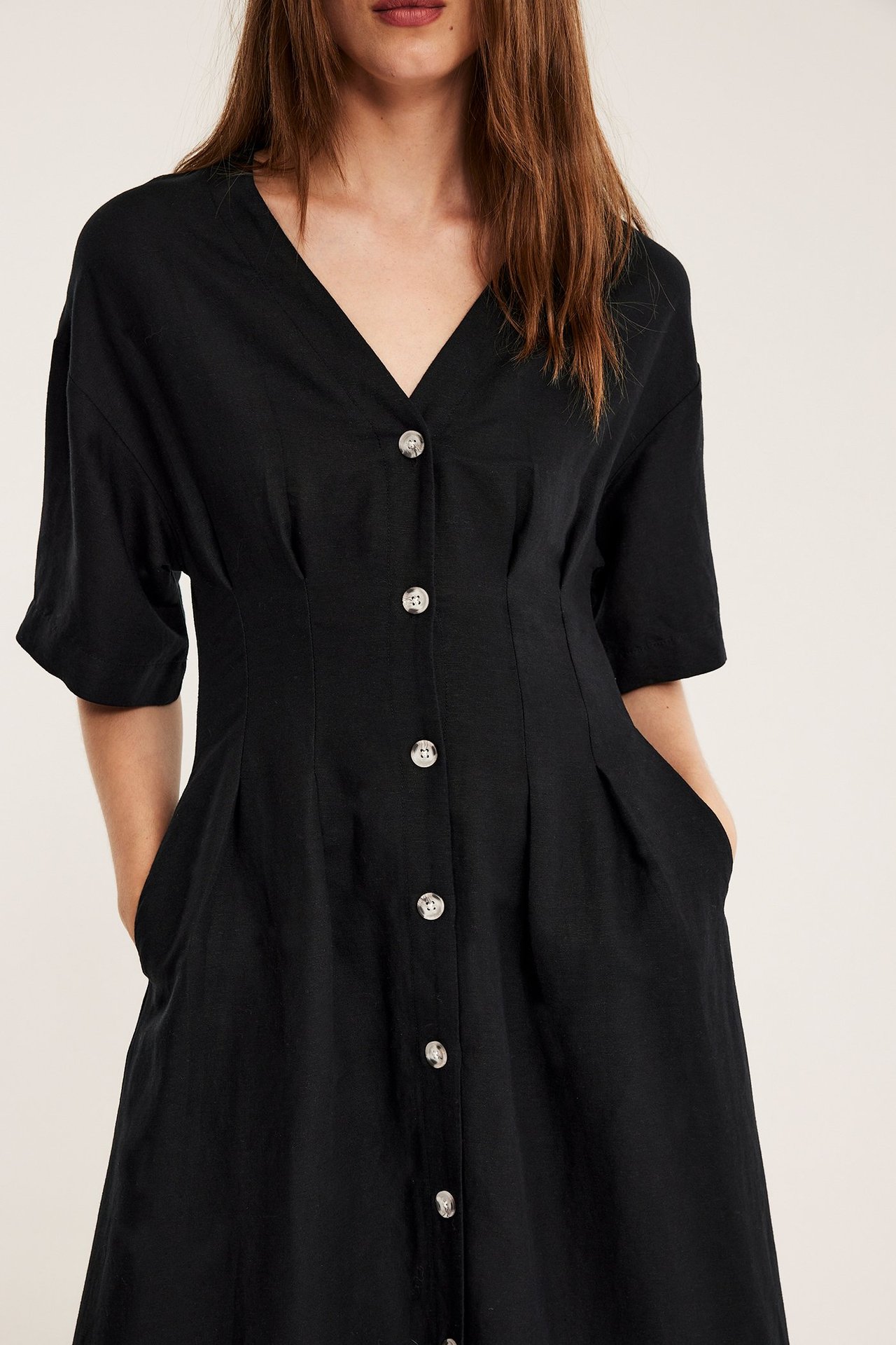 Edestä: Nainen mustassa pellavasekoitemekossa, jossa V-pääntie, etunapit ja kädet taskuissa.