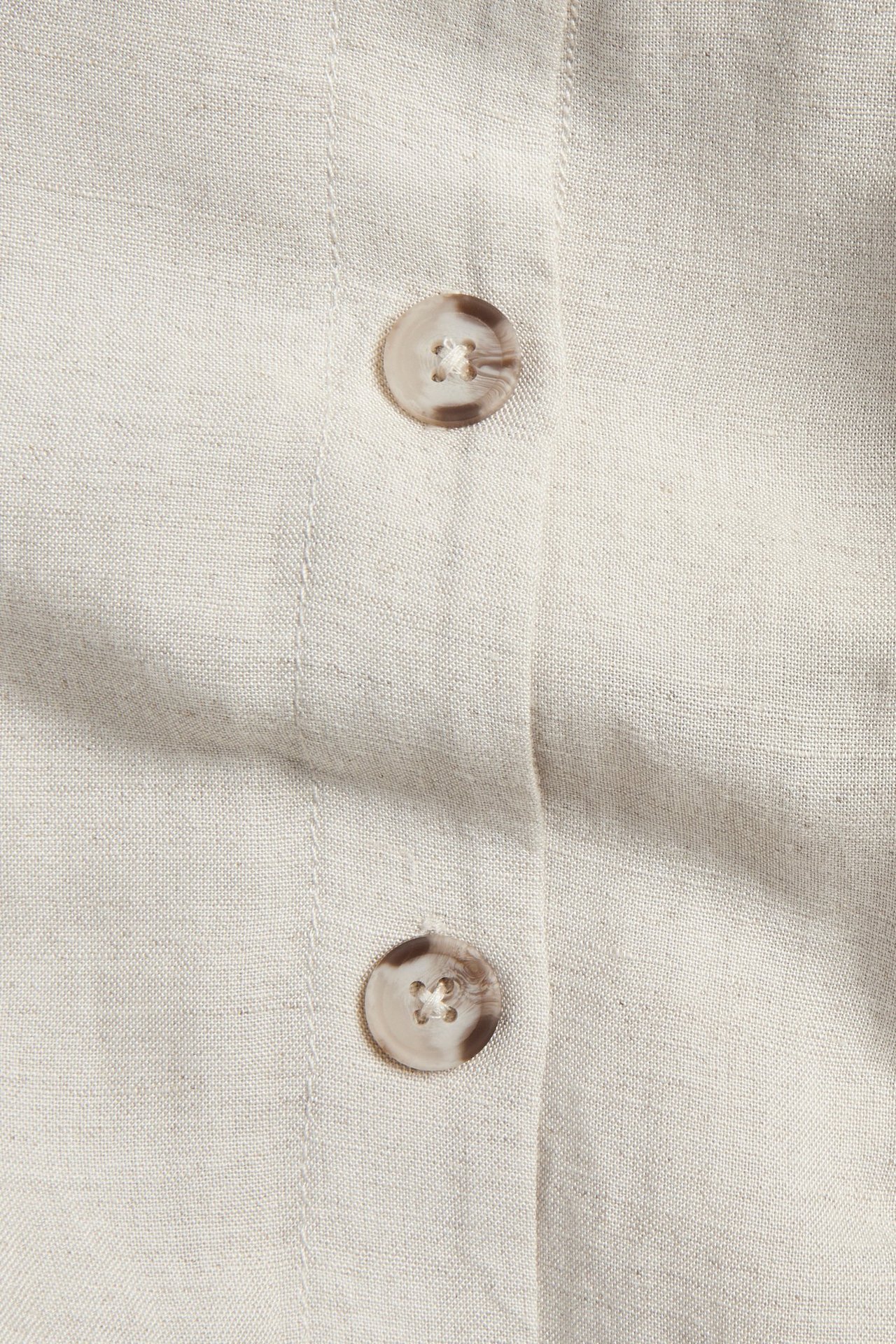 Zbliżenie beżowej sukienki z mieszanki lnu z dwoma okrągłymi guzikami w wirujący wzór z przodu.