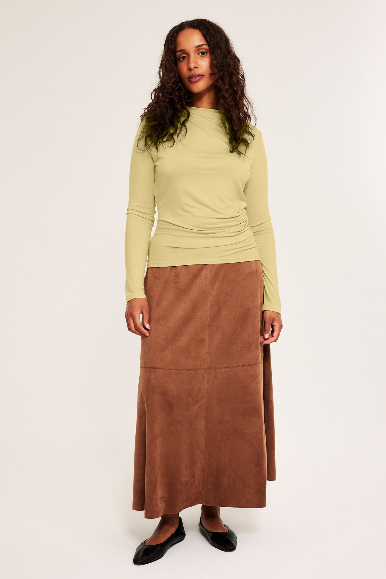Frontvisning av en kvinne i lys gul drapert viskosetopp, brunt skjørt og svarte flate sko.