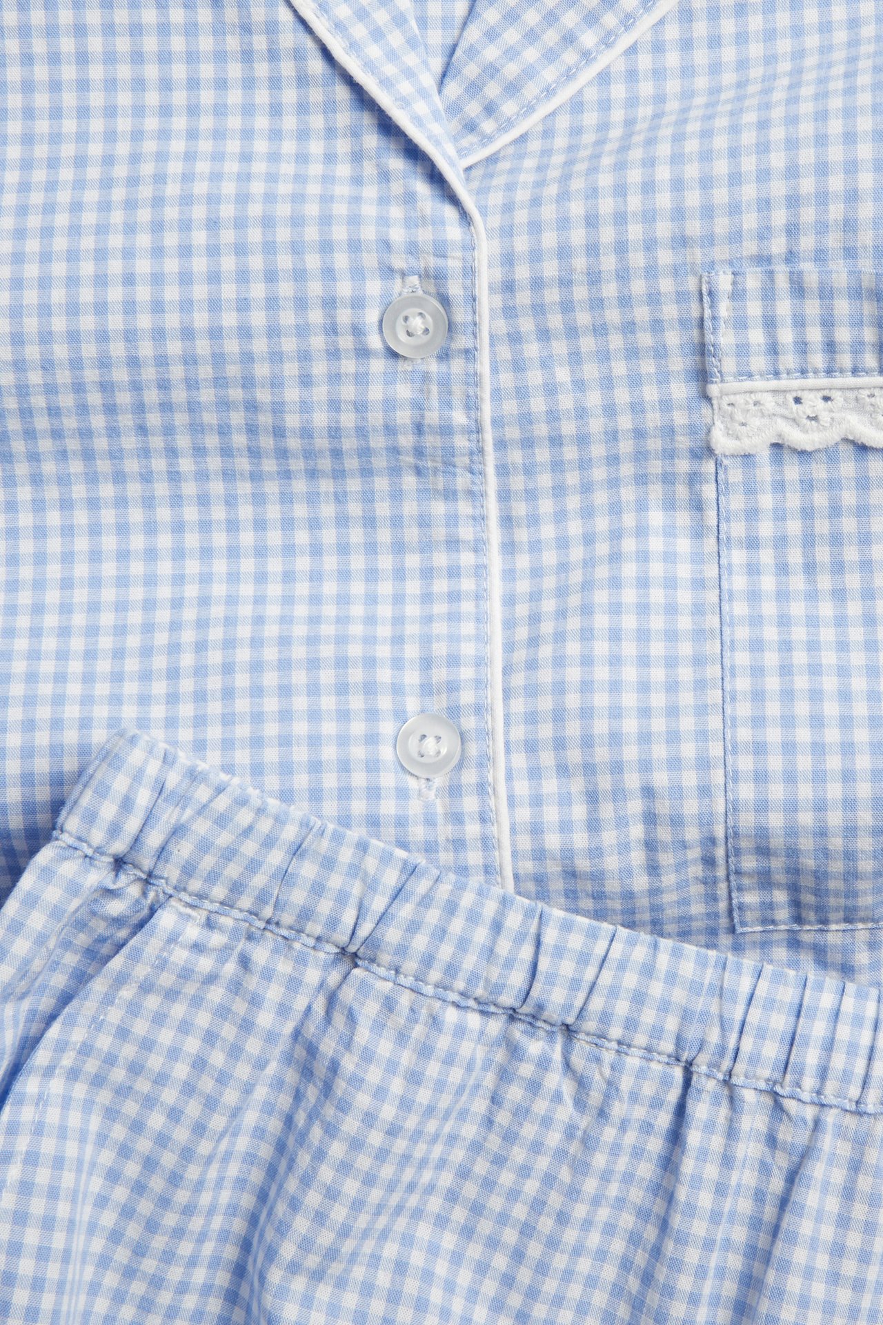 Nærbilde av blå- og hvitrutet pyjamas i bomullspoplin med hvite passpoaler og blondekantet brystlomme.