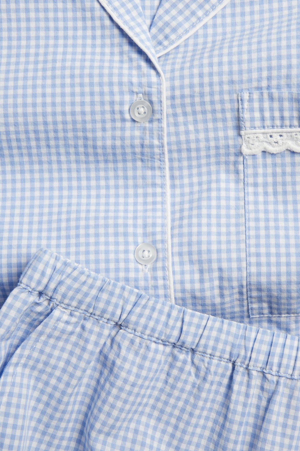 Närbild av blåvitrutig pyjamas i bomullspoplin med vit passpoal och spetskantad bröstficka.