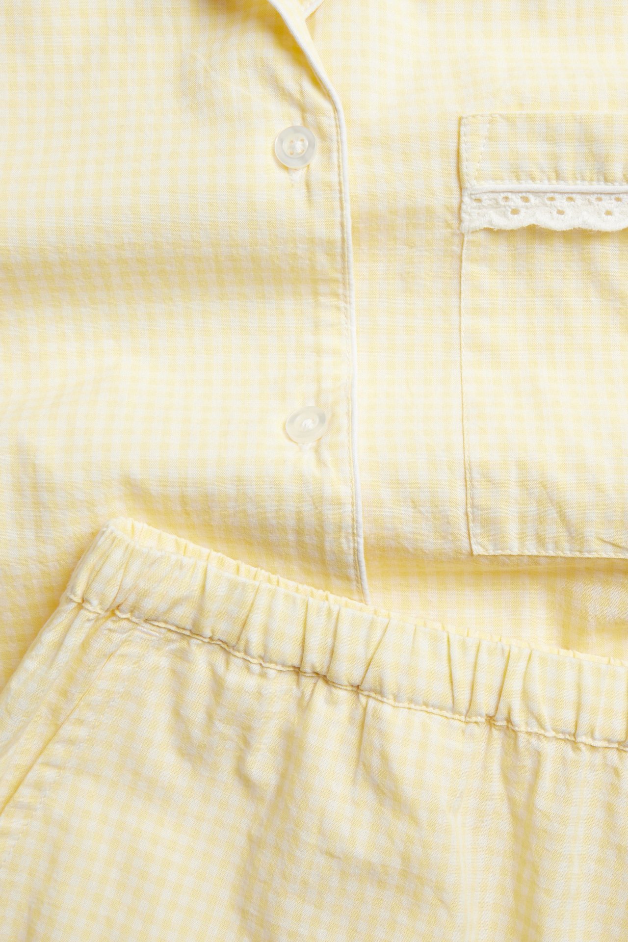Nærbilde av et lys beige rutete bomullspyjamassett med hvite passpoaler, blondelomme og elastiske shorts.