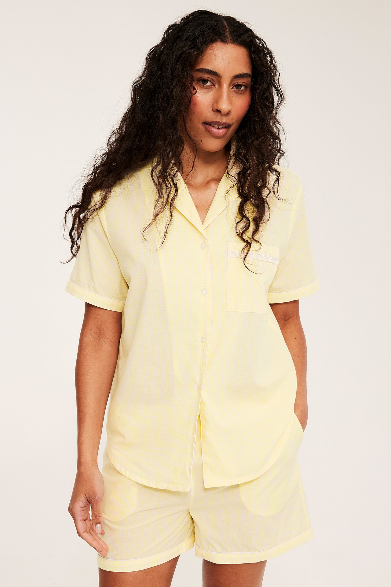 Edestä: Nainen vaaleankeltaisessa pikkuruudullisessa puuvillapopliinipyjamasetissä lyhythihaisella paidalla ja shortseilla.