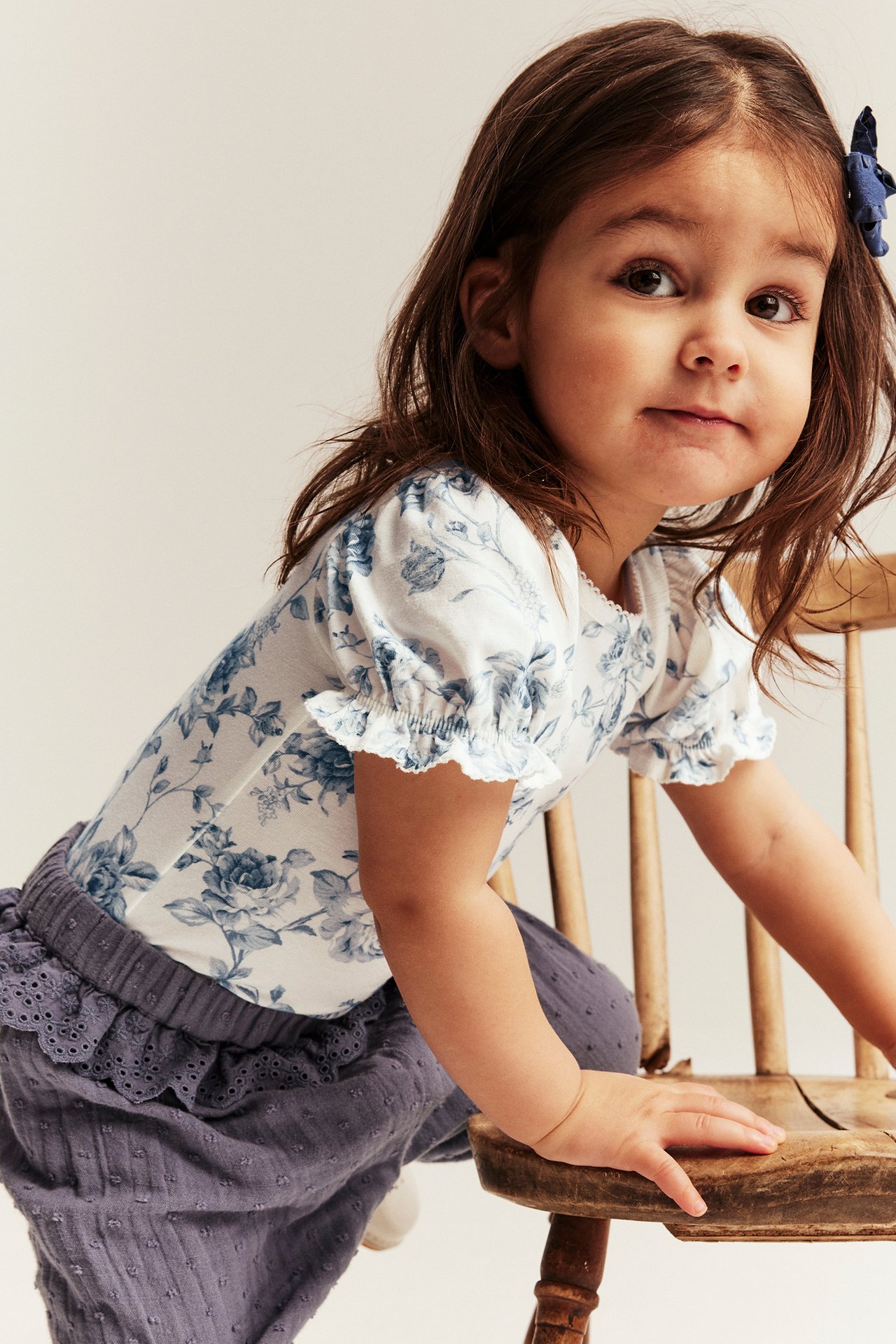 Framifrån, ett barn i vitblommig kortärmad body, mörkgrå byxor och ett blått hårspänne.