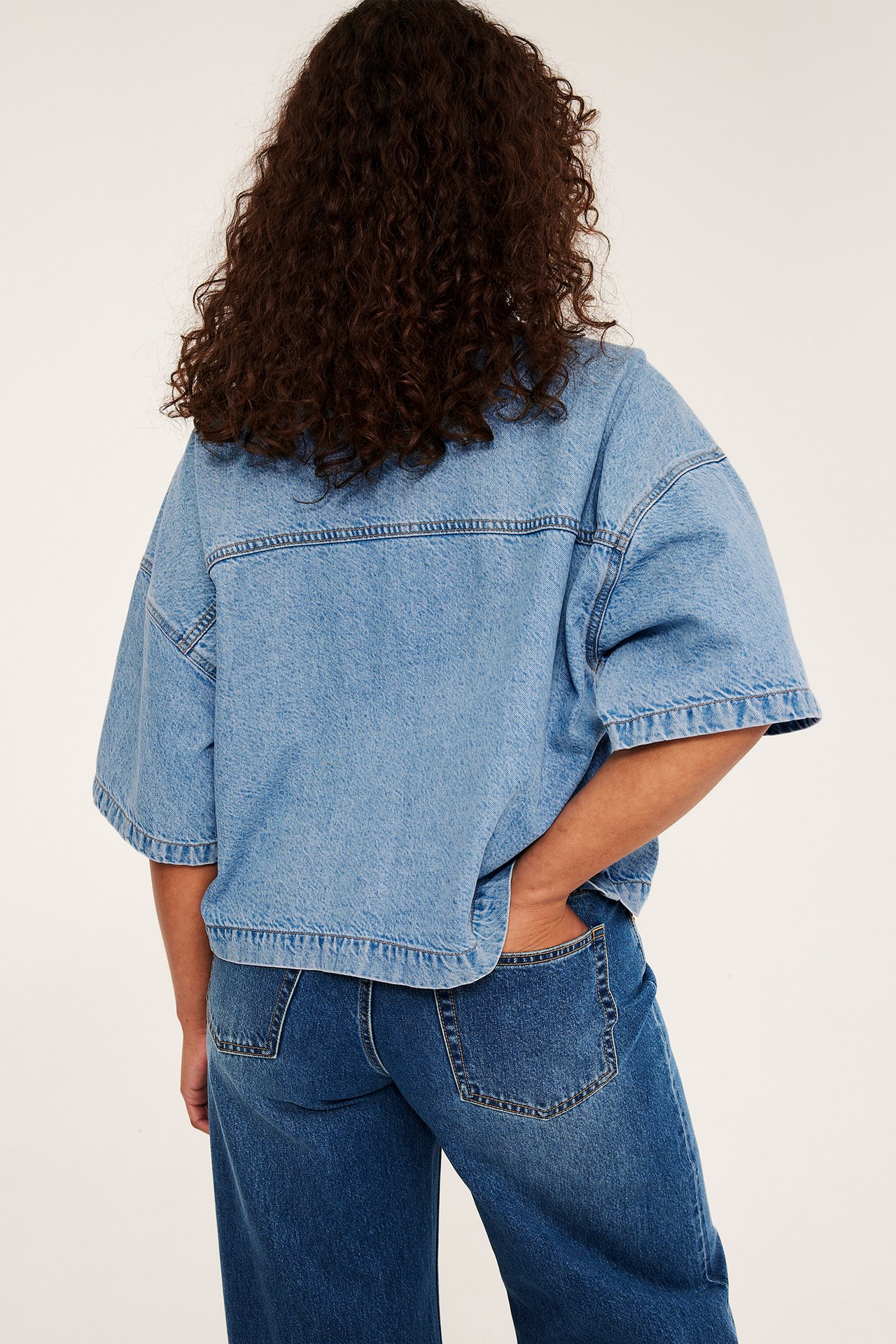Bakfra: kvinne i en lys blå, kort, boxy denimskjorte med korte ermer og mørke jeans.