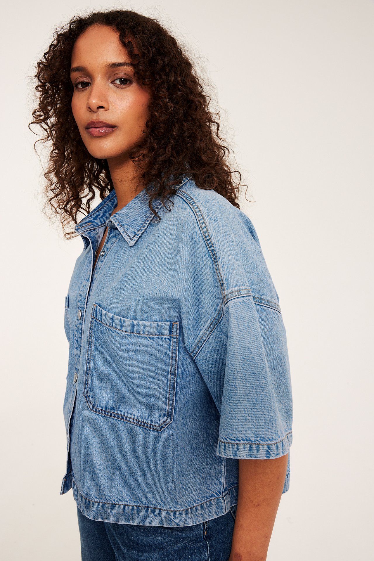 Nærbilde av en kvinne i en lyseblå, boxy, kortermet croppet dongeriskjorte.