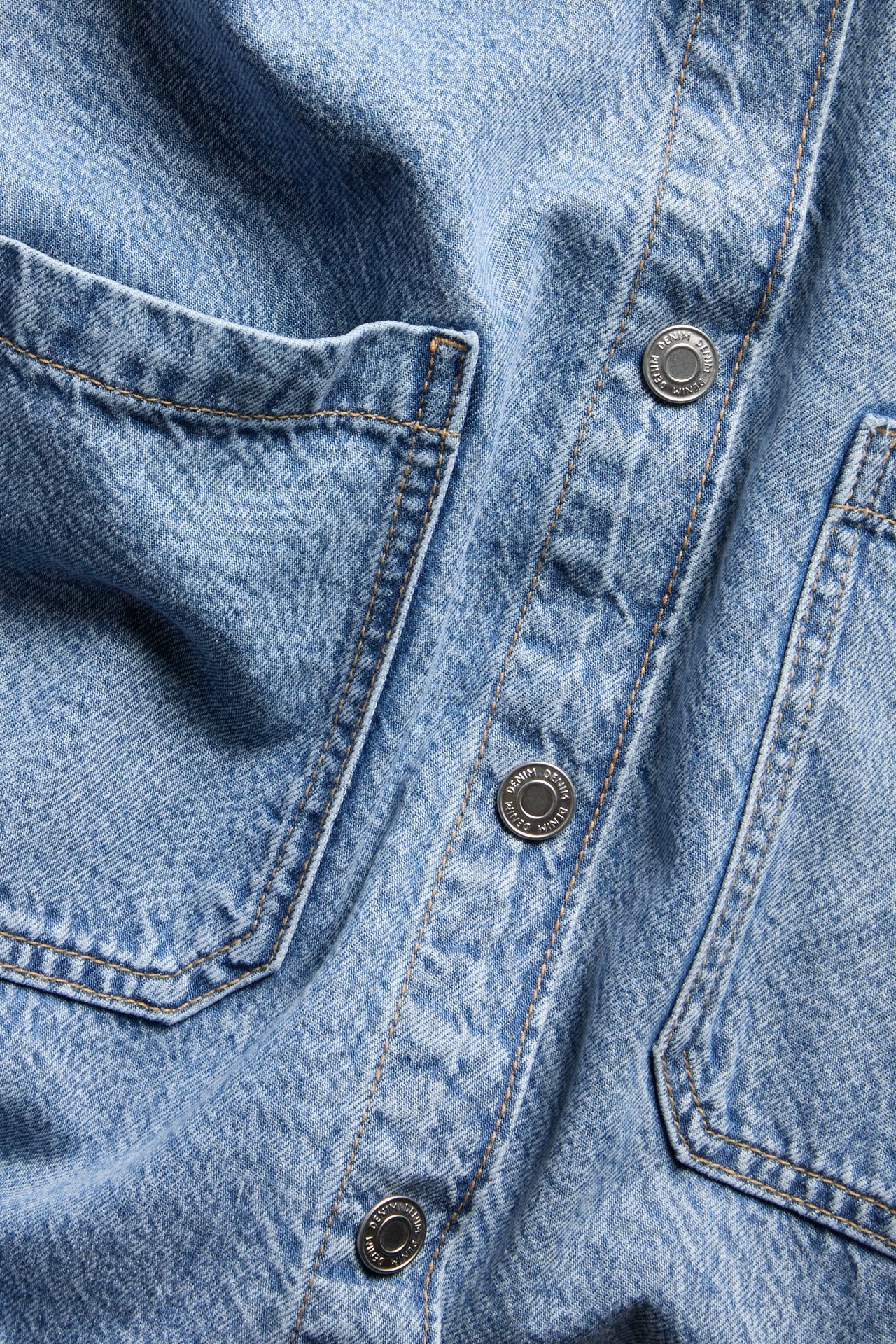 Lähikuva edestä vaaleansinisestä, lyhythihaisesta, laatikkomallisesta farkkupaidasta, jossa taskut ja hopeanapit.