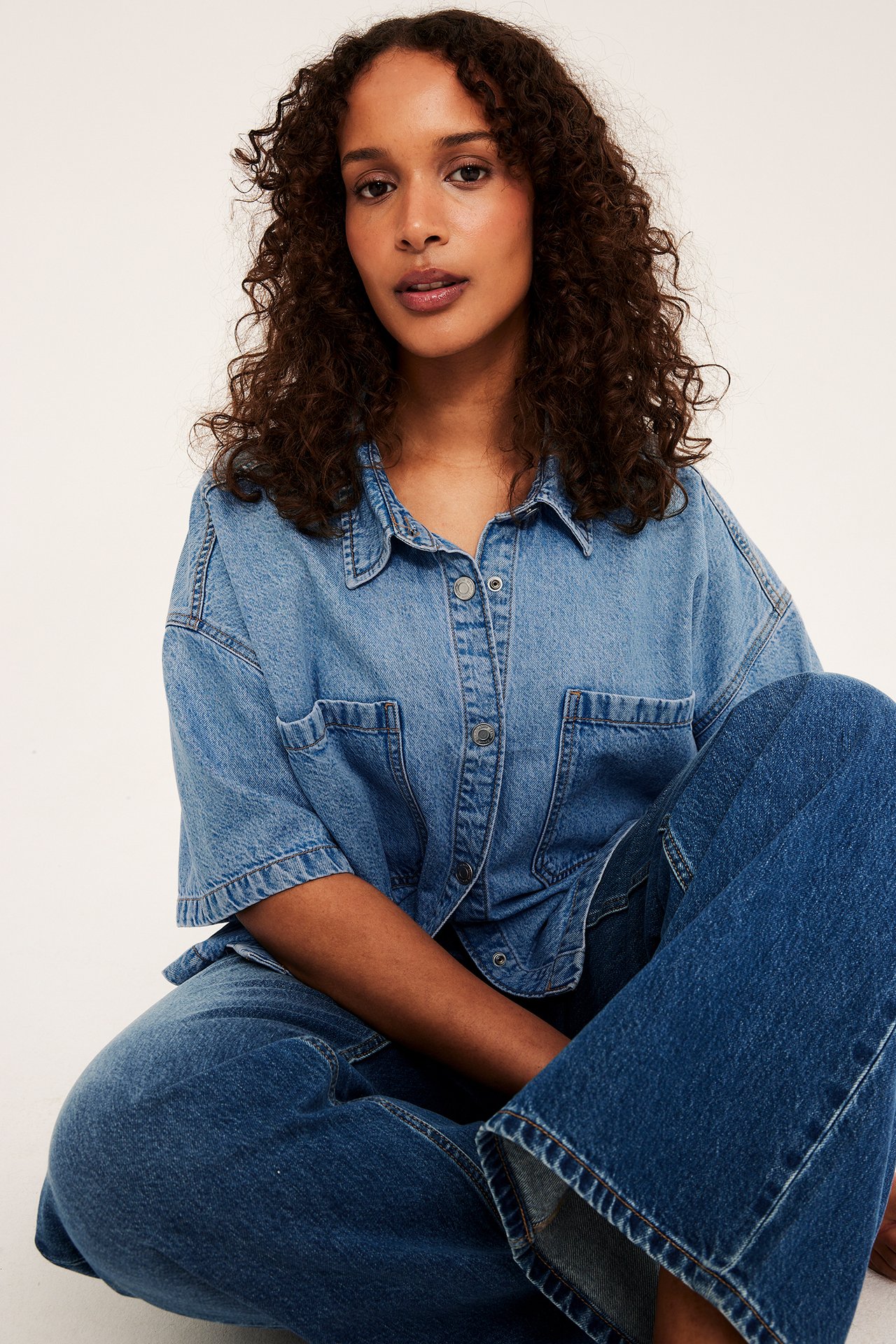 Frontvy av kvinna i ljusblå, boxig jeansskjorta med kort ärm och mörkblå vida jeans.
