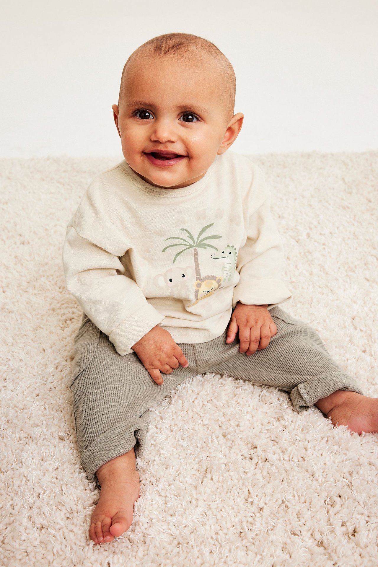 Framifrån: en baby i ljusbeige bomulls-t-shirt med djurtryck och grå ribbade byxor.