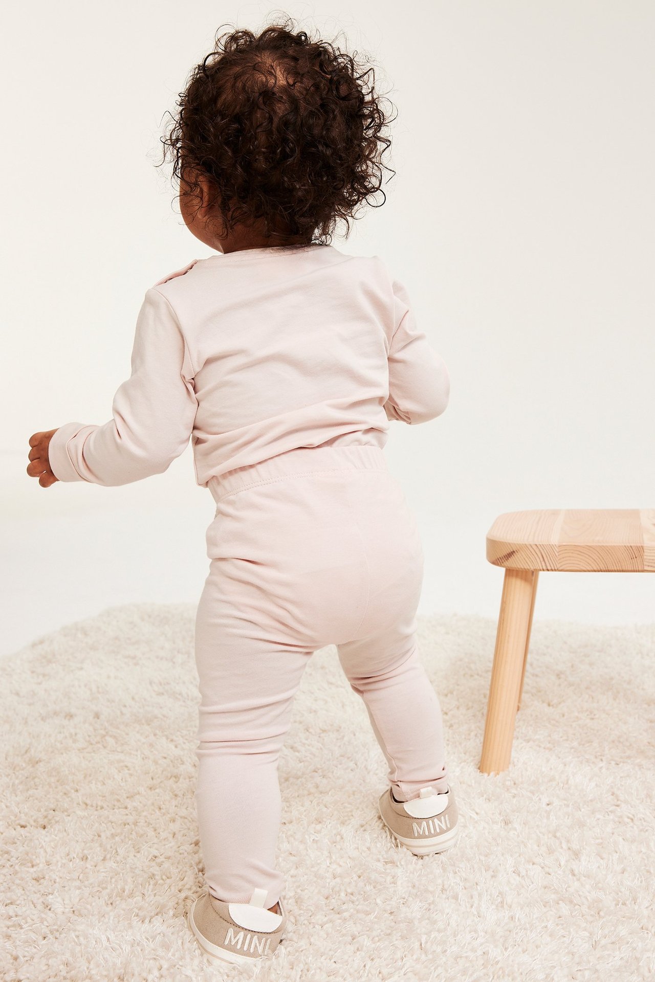 Vauva takaa: vaaleanpunaiset tyköistuvat leggingsit, vaaleanpunainen pitkähihainen paita ja beiget vauvakengät.