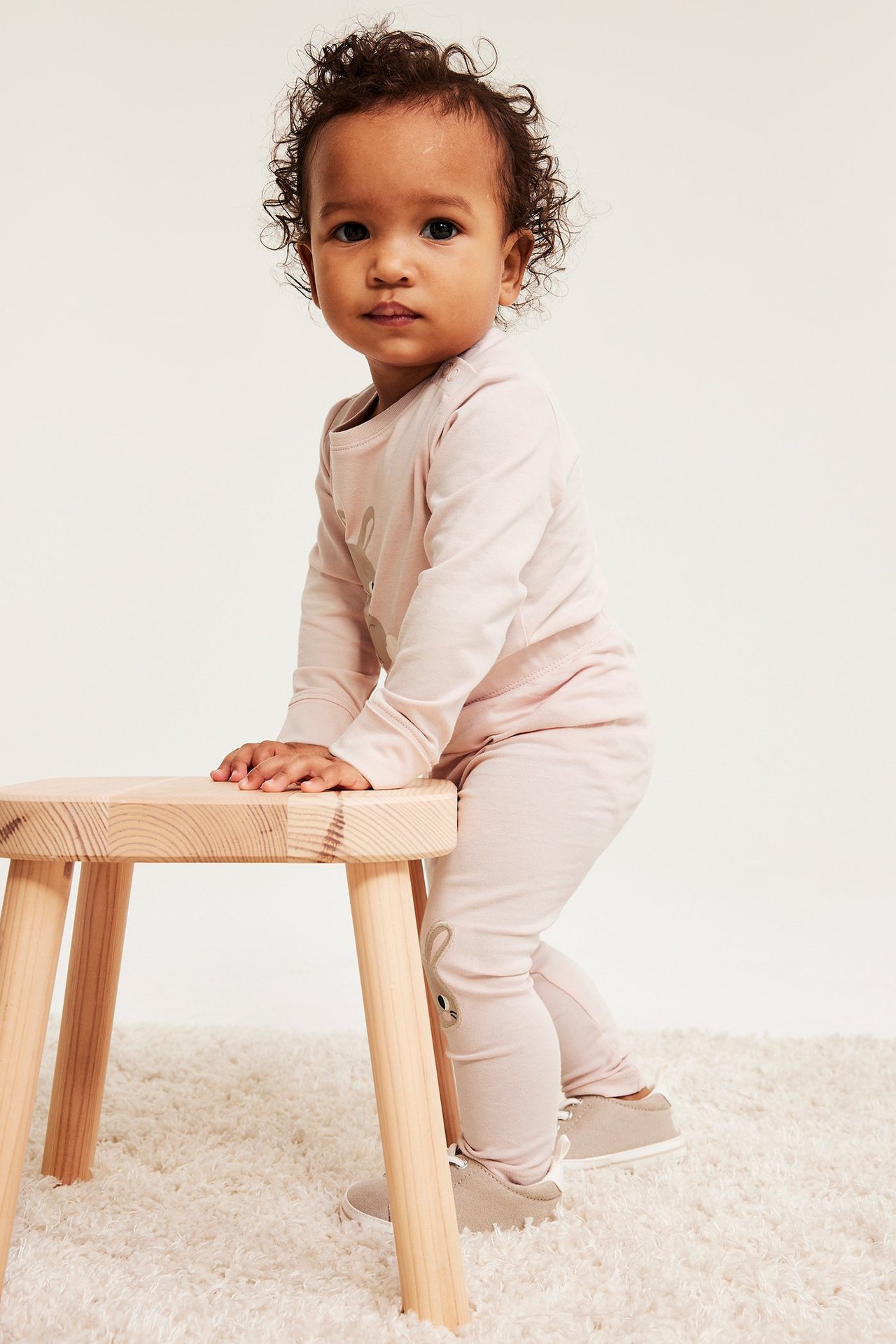 Edestäpäin: Vauva vaaleanpunaisissa leggingseissä, joissa pupuapplikaatiot, yhteensopivassa paidassa ja beigenvärisissä kengissä.