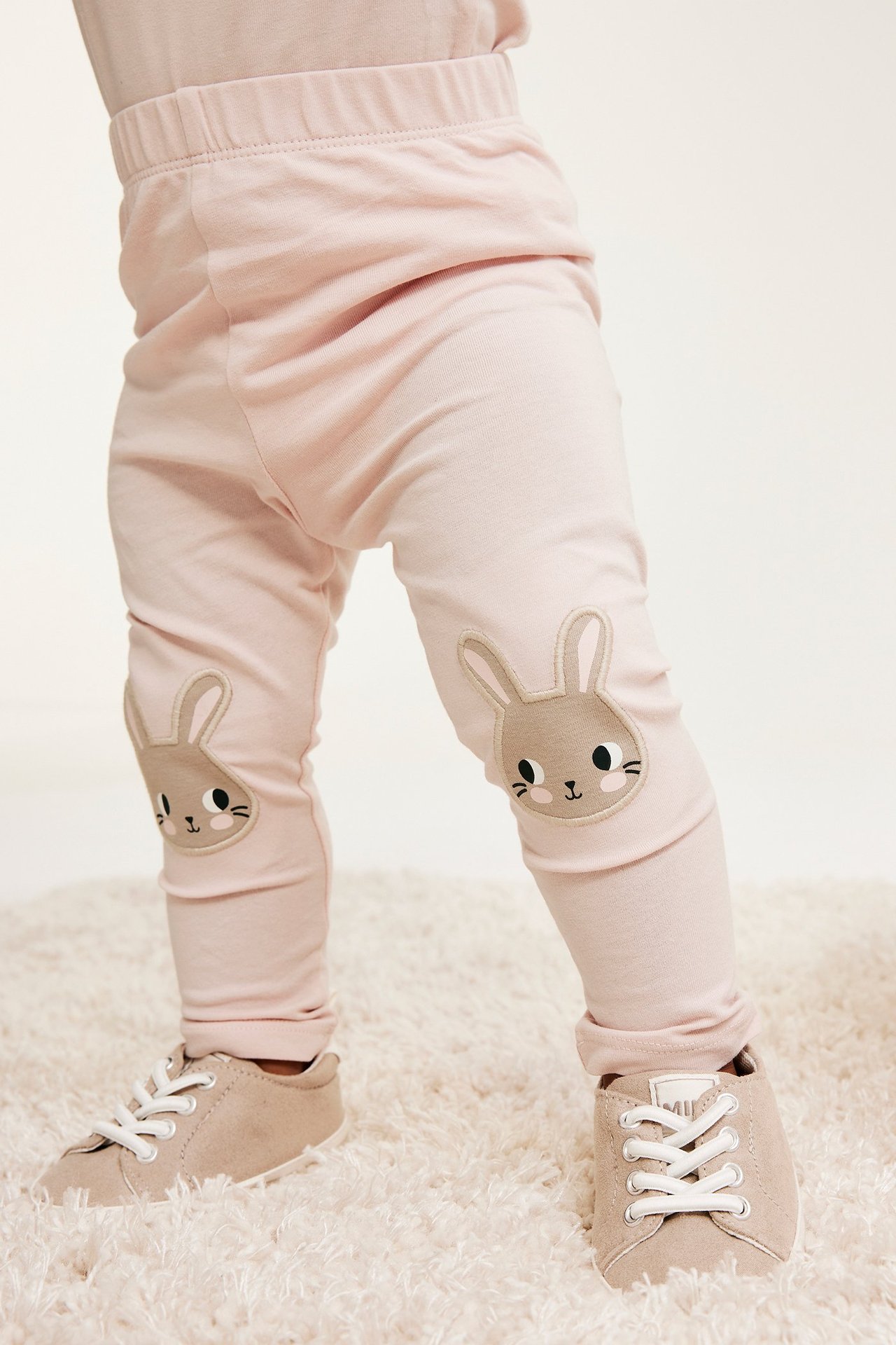 Edestä päin, vauvan jalat vaaleanpunaisissa leggingseissä pupuapplikaatioilla polvissa ja beiget nauhakengät.