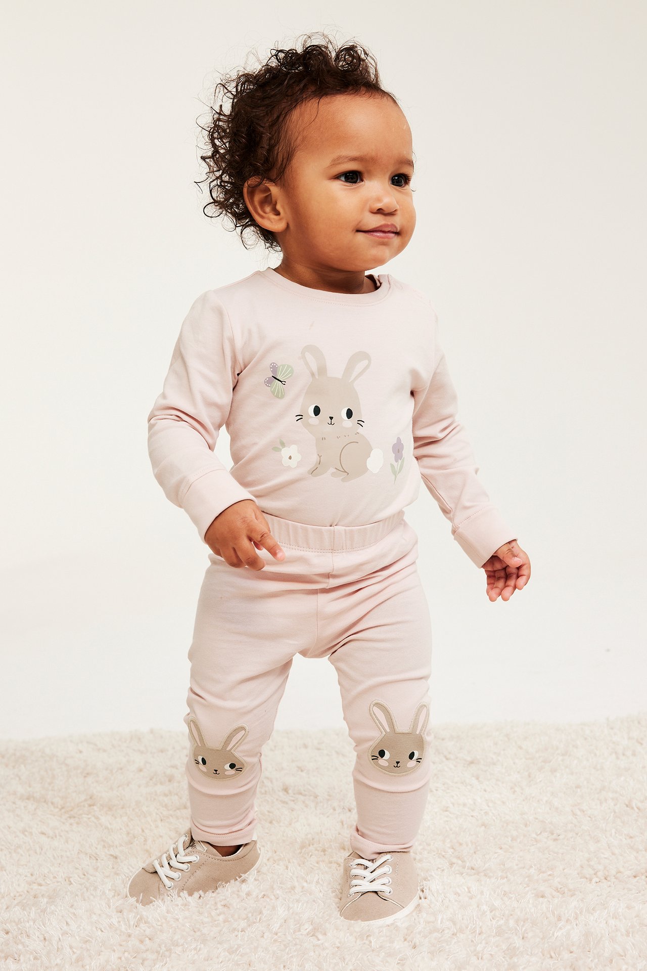 Edestäpäin, vauva yllään vaaleanpunaiset legginsit pupuapplikaatioilla polvissa, yhteensopiva toppi ja beiget kengät.