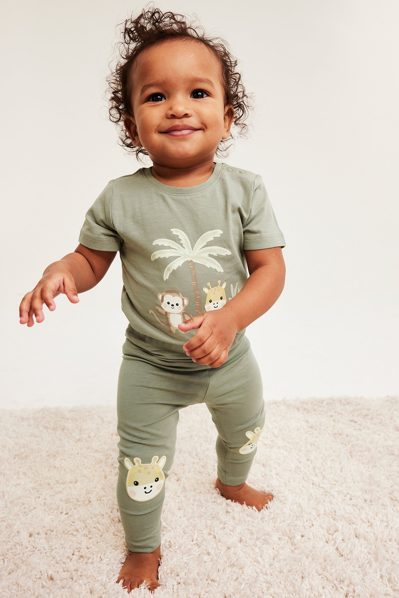 Framifrån: leende baby i mörkgröna leggings med giraffapplikationer på knäna och matchande mönstrad t-shirt.