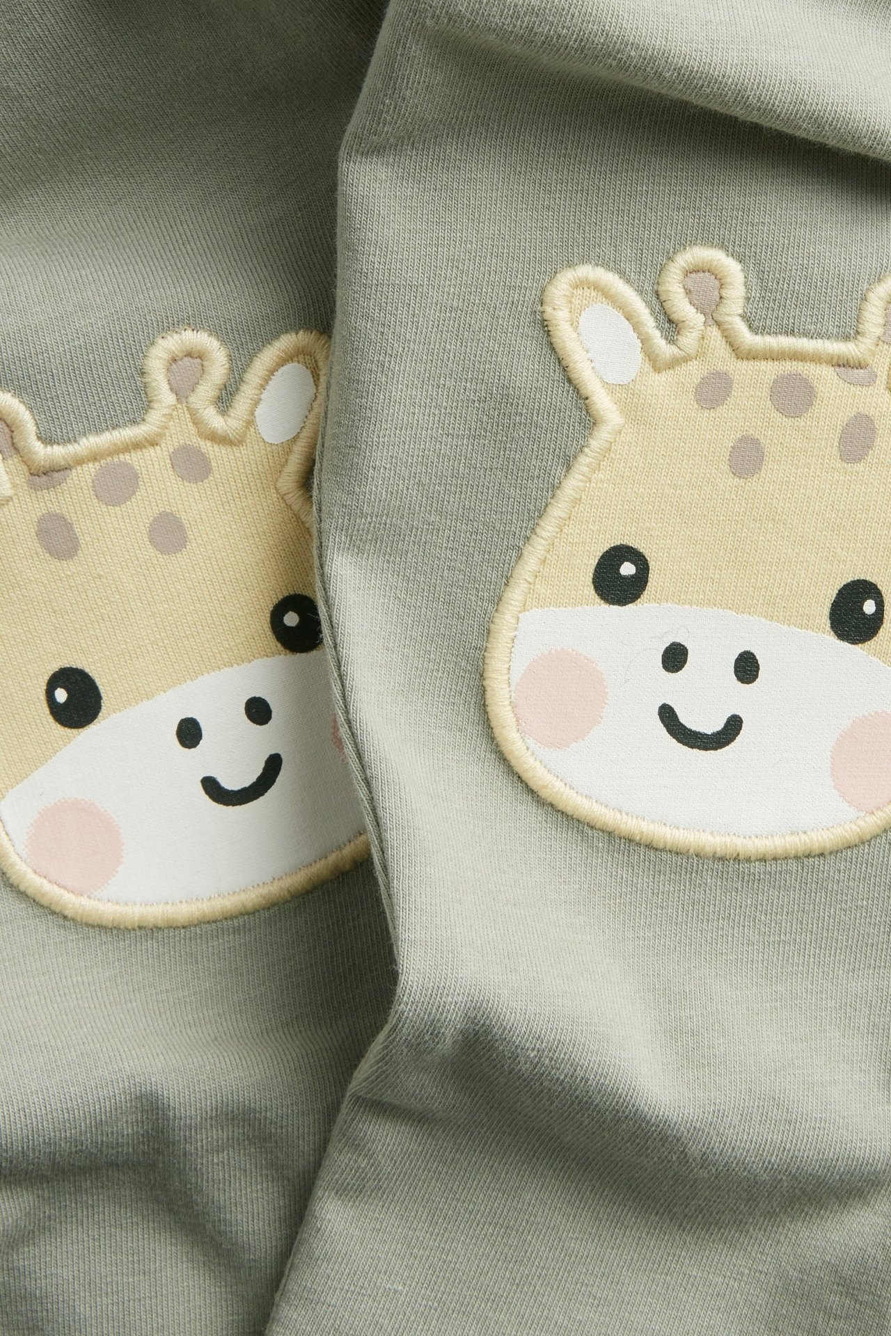 Zbliżenie na ciemnozielone legginsy niemowlęce z jasnobeżową aplikacją głowy żyrafy na każdym kolanie.