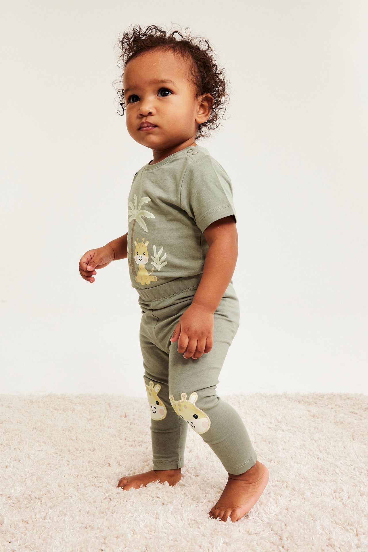 Sidovy av en baby i mörkgröna bomullsleggings med giraffapplikationer på knäna och matchande t-shirt.