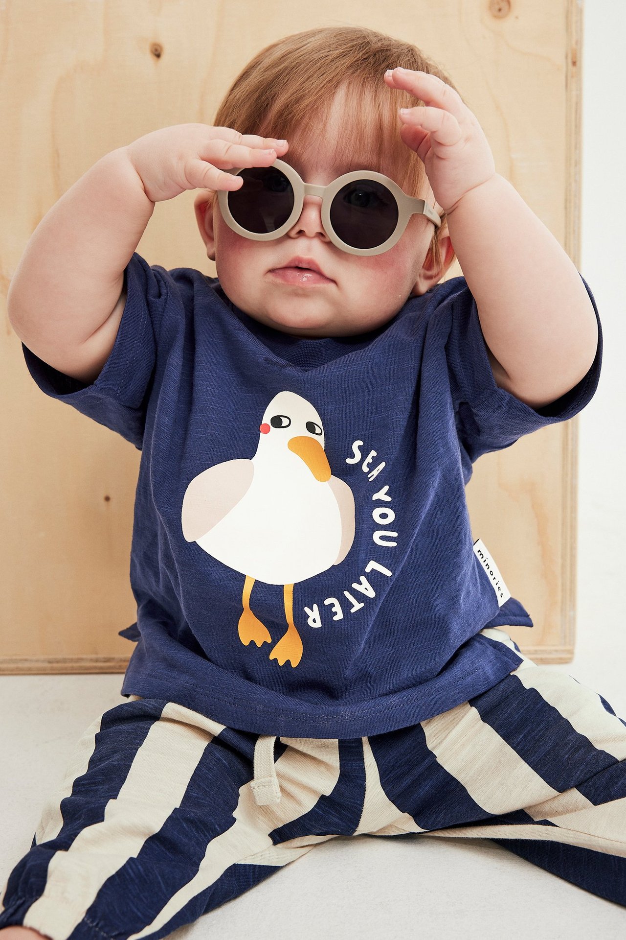 Framsida av baby i blå bomulls-T-shirt med måstryck, randiga joggingbyxor och beige solglasögon.