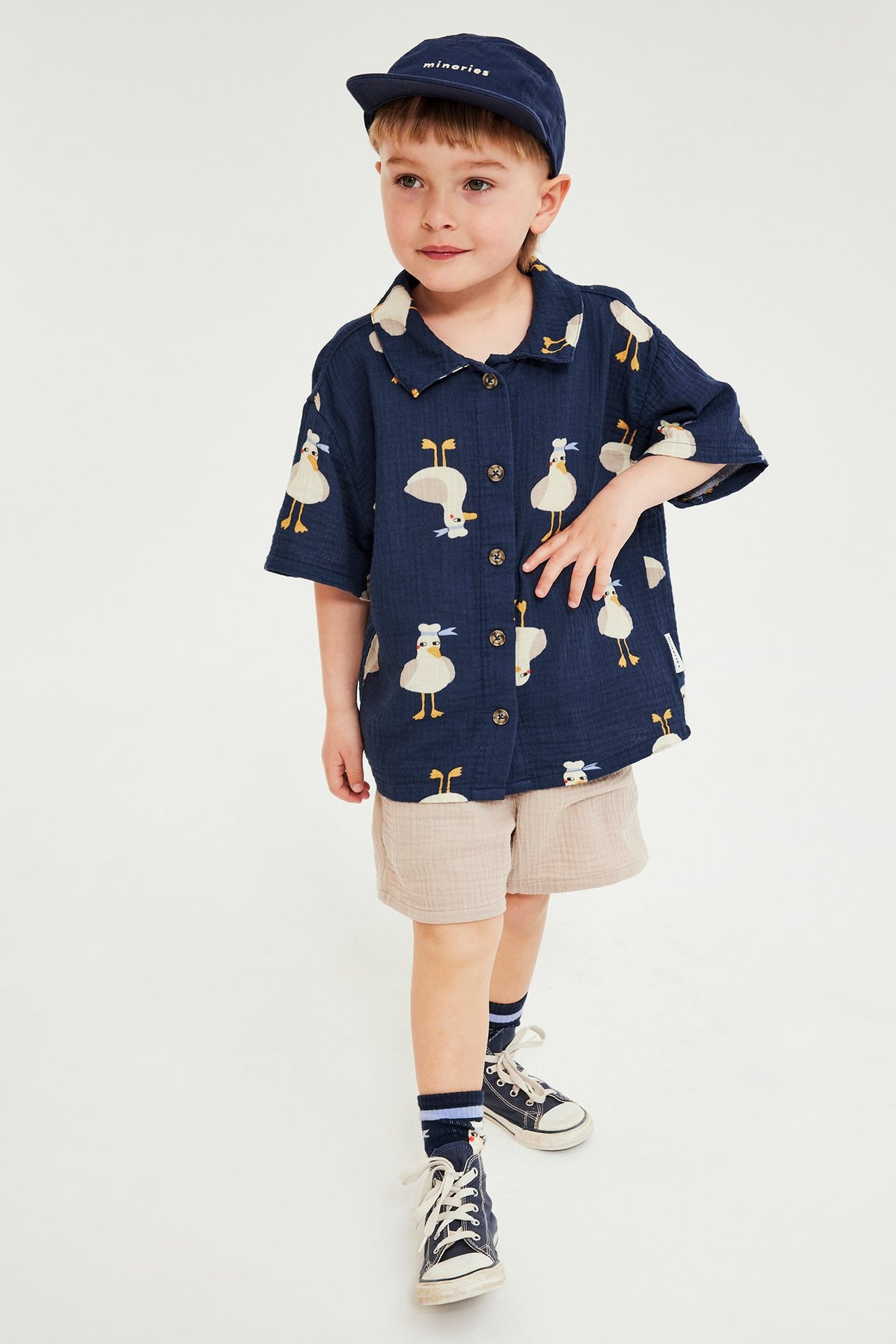 Framifrån, barn i blå mönstrad kortärmad musslins-skjorta, beige shorts, blå keps och sneakers.