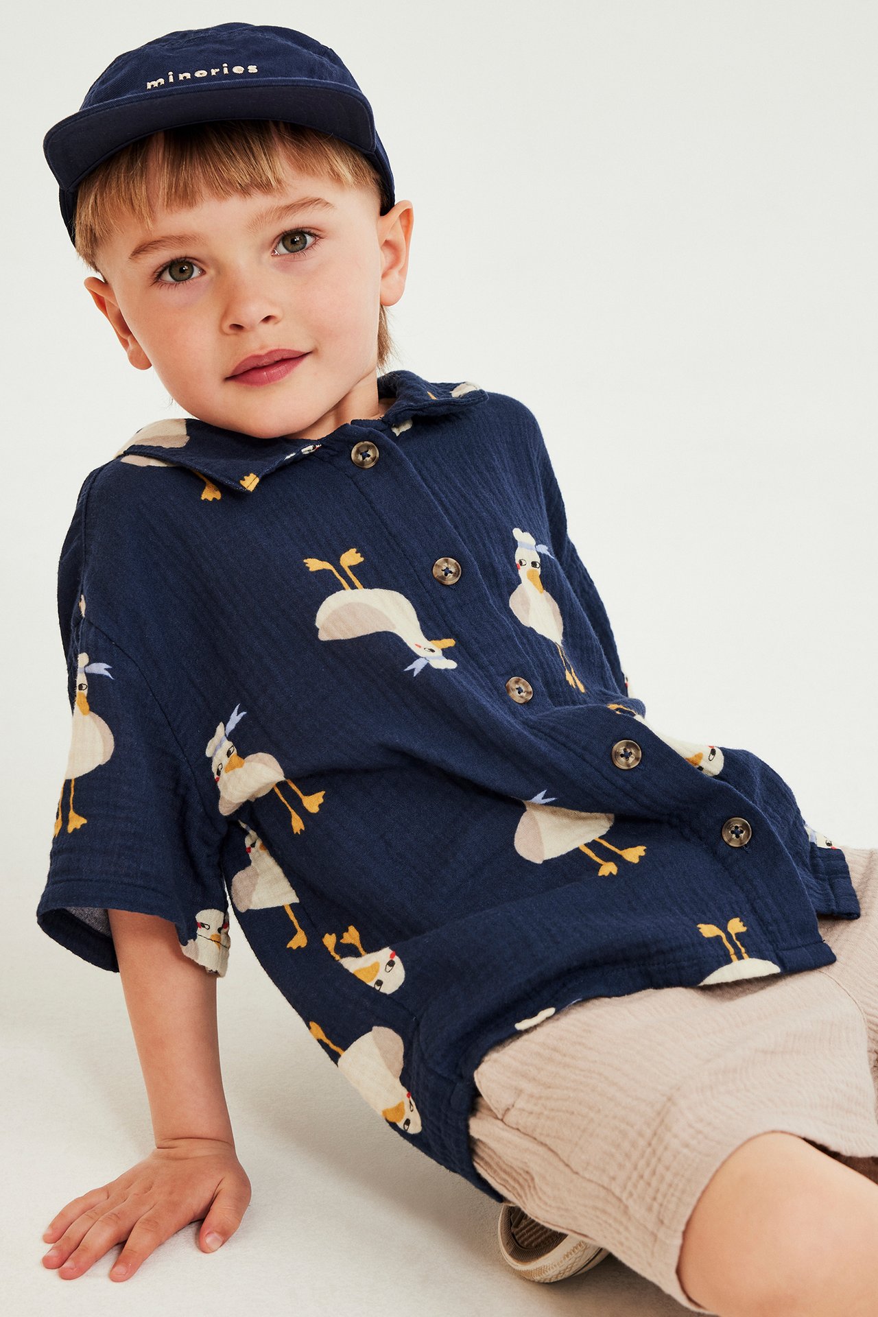 Front view av et barn i blå ande-mønstret kortermet musselinskjorte, lysebeige shorts og marineblå caps.