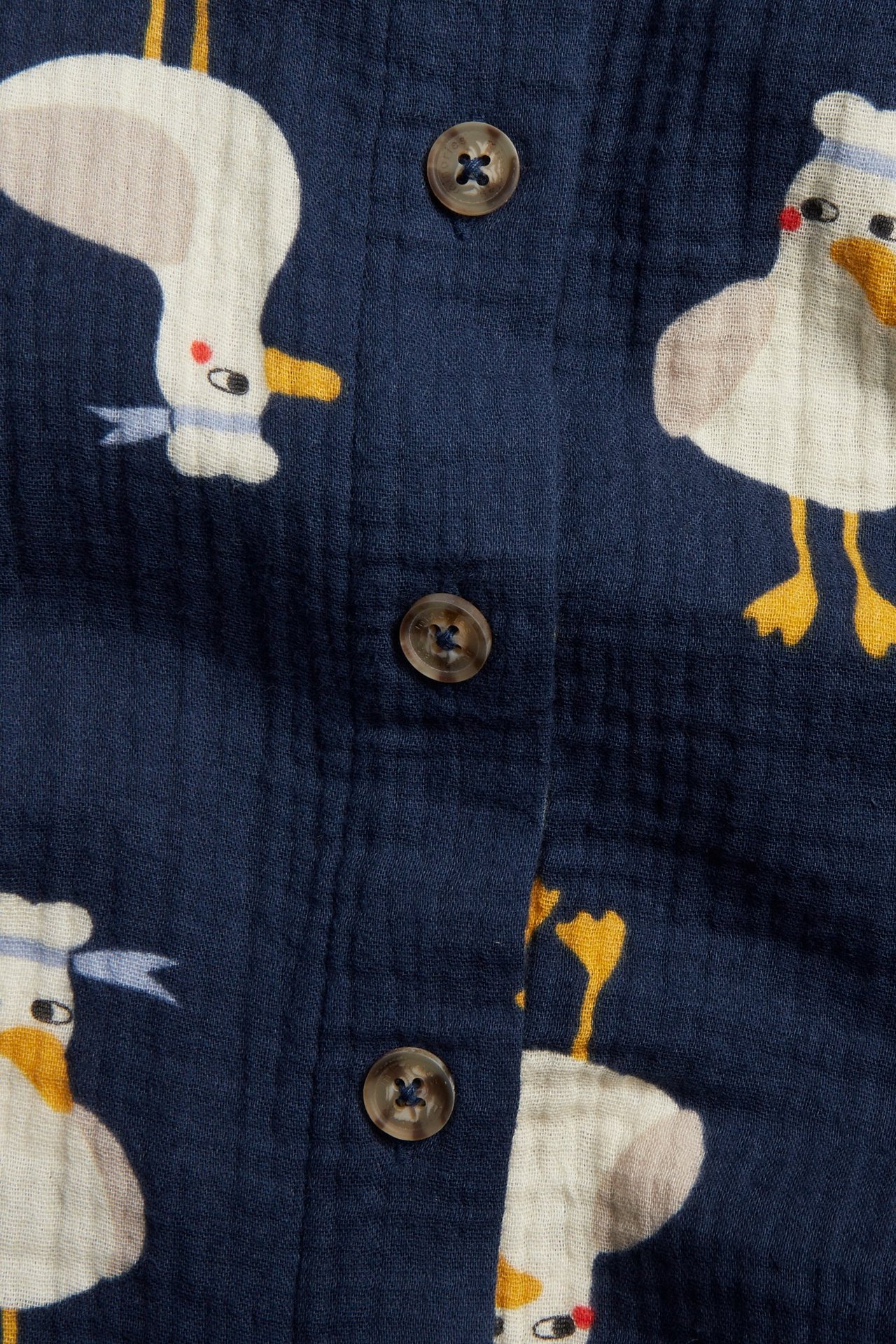 Nærbilde av en blå kortermet muslin-skjorte for barn med måkemønster og brune knapper.