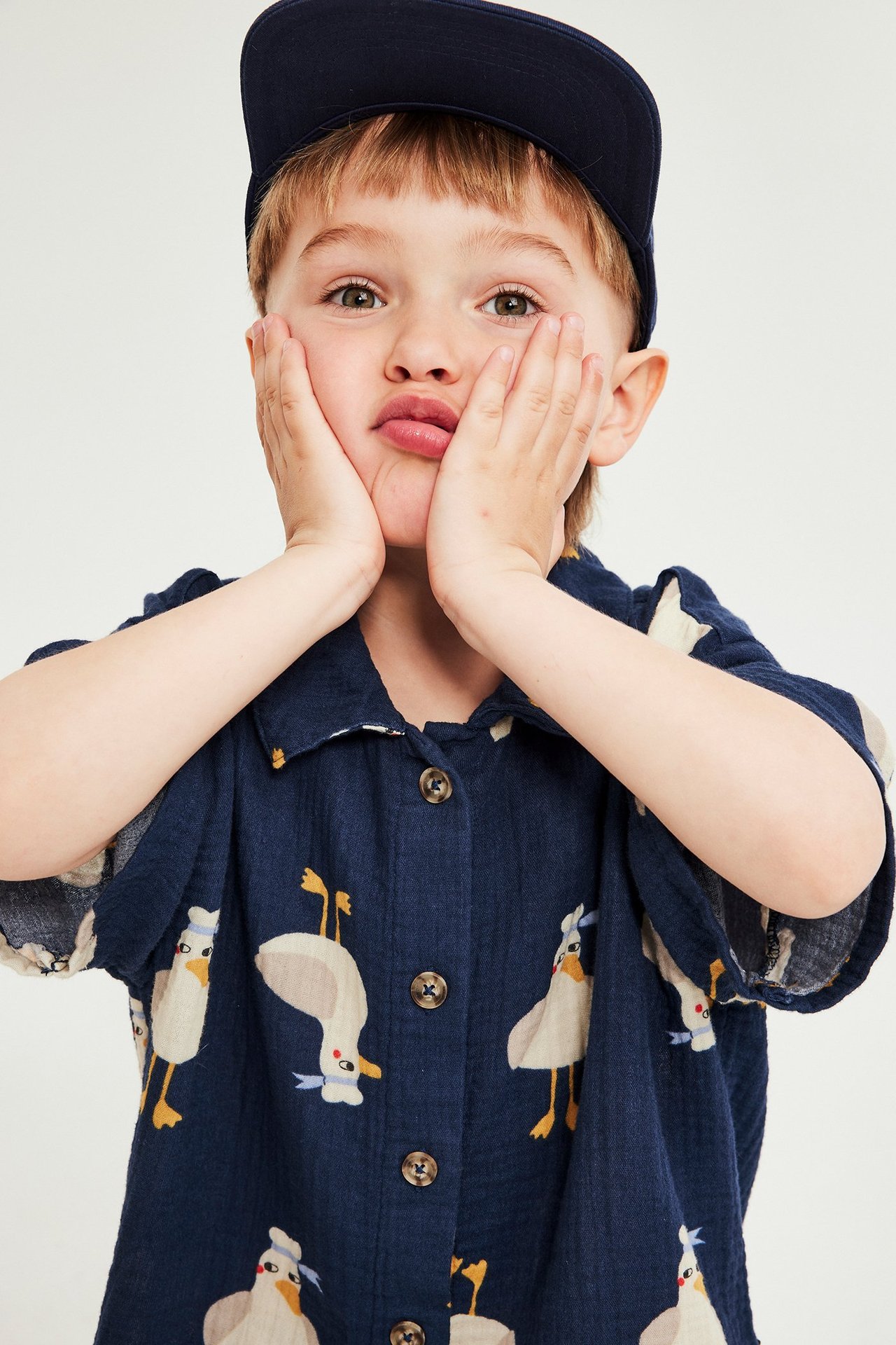 Barn bär en marinblå mönstrad kortärmad muslinskjorta med knappar och en marinblå keps.