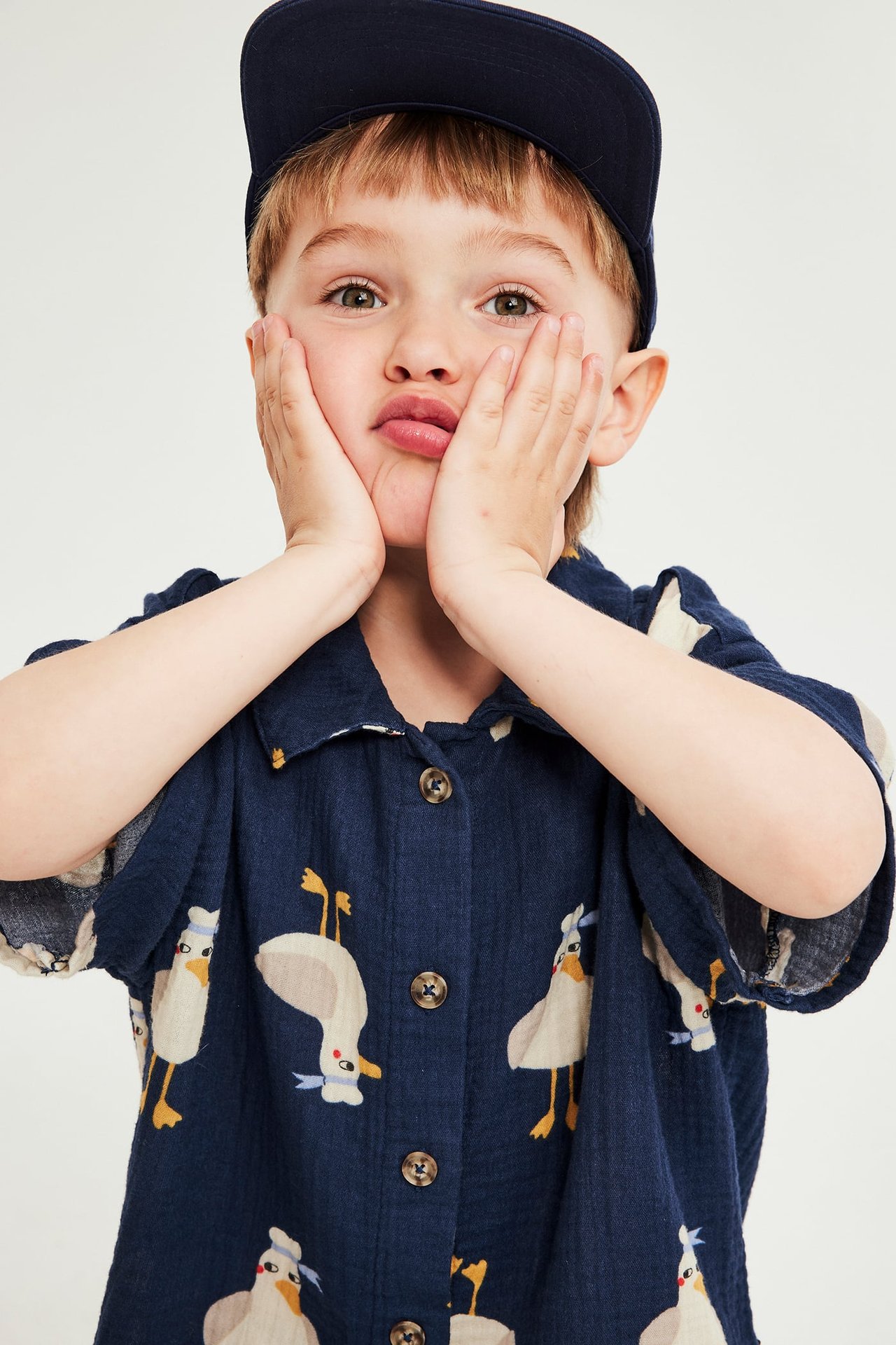 Barn bär en marinblå mönstrad kortärmad muslinskjorta med knappar och en marinblå keps.