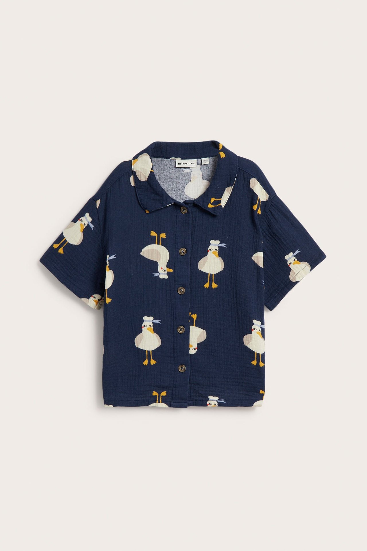 Framifrån, en marinblå kortärmad musslinskjorta för barn med vitt och gult ankmönster och knappar.