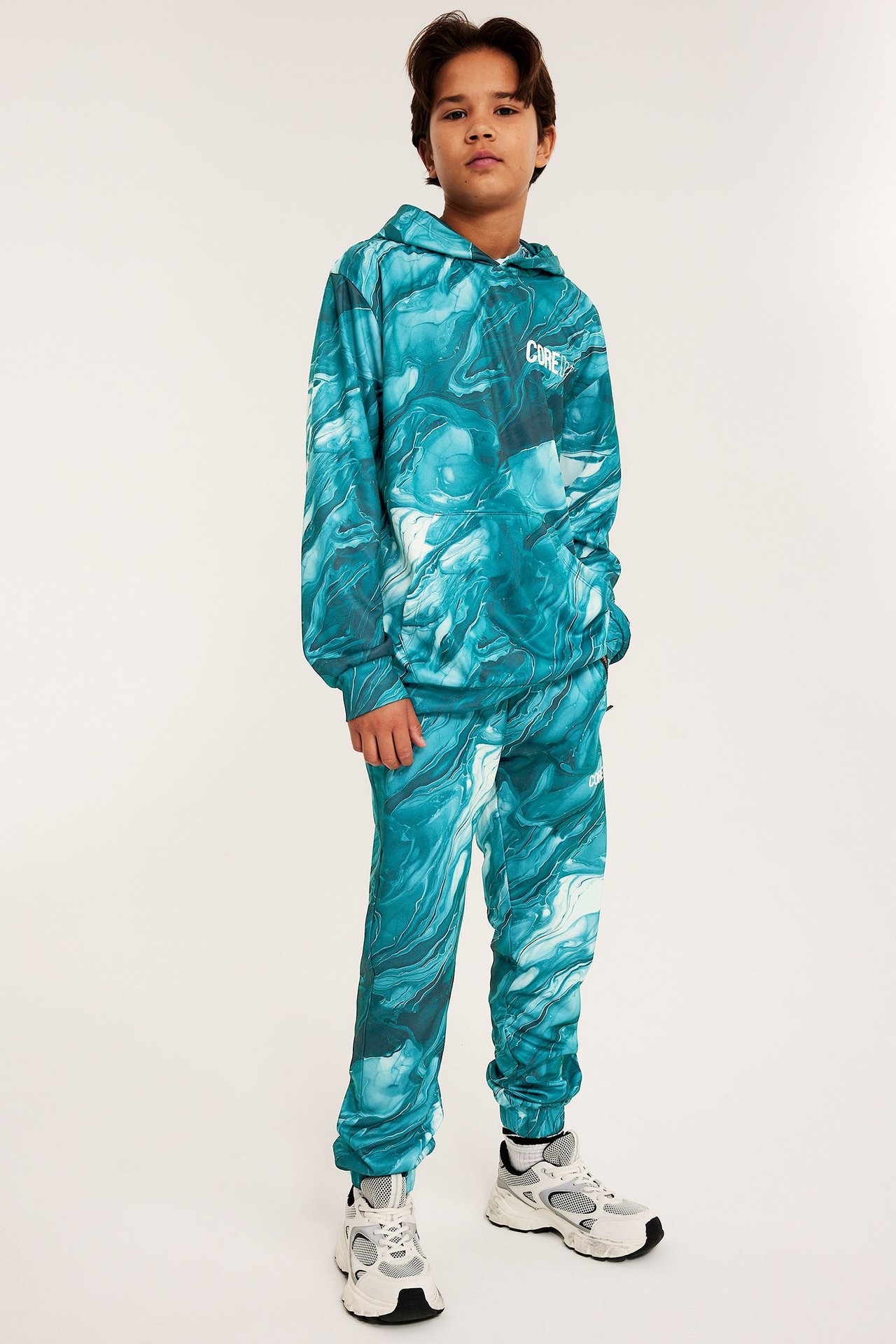 Widok z przodu: Dziecko w zielonej, abstrakcyjnej bluzie WCT z kieszenią i pasujących joggerach.