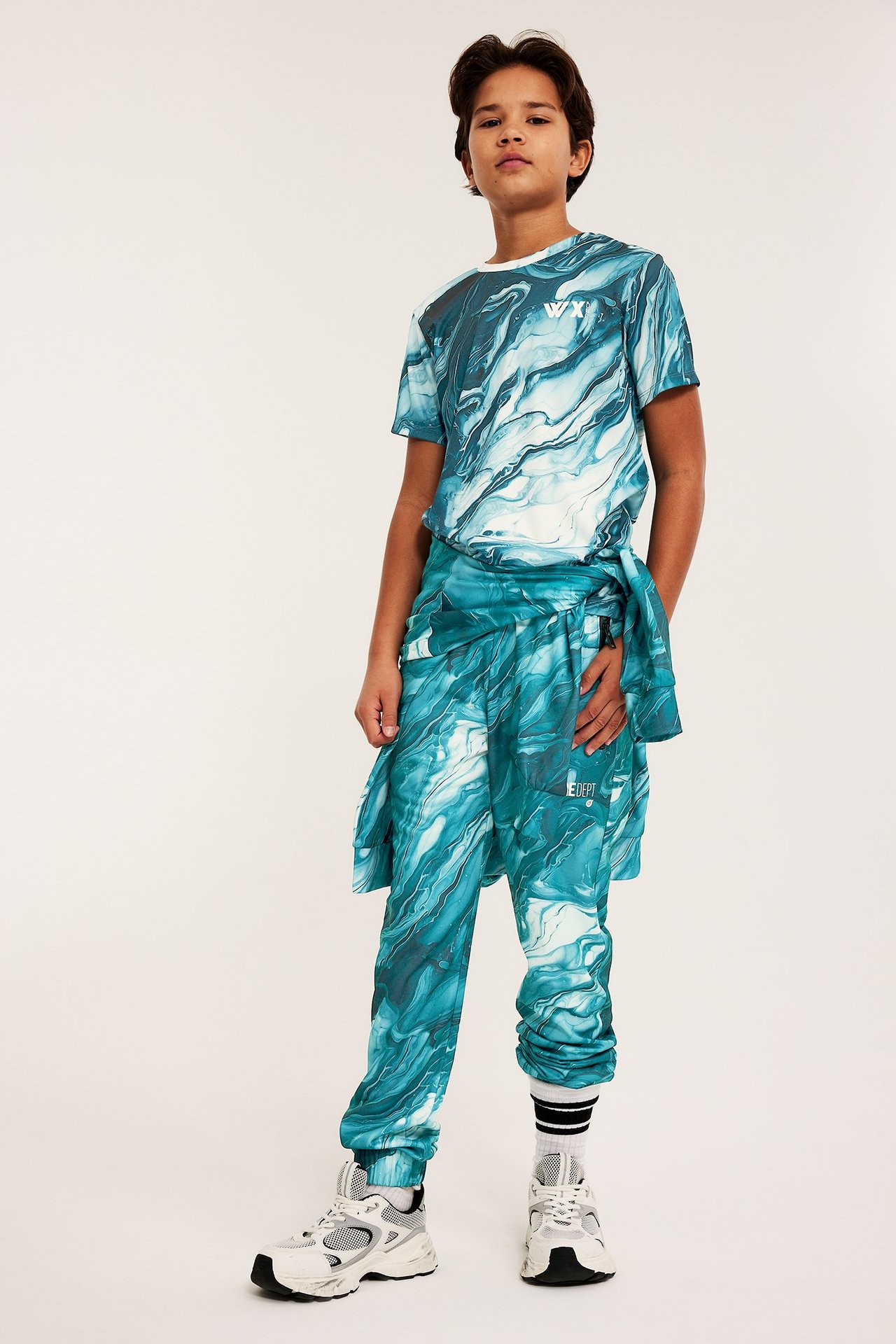 Widok z przodu dziecka w zielonych, wzorzystych spodniach dresowych, pasującym T-shircie i białych sneakersach.