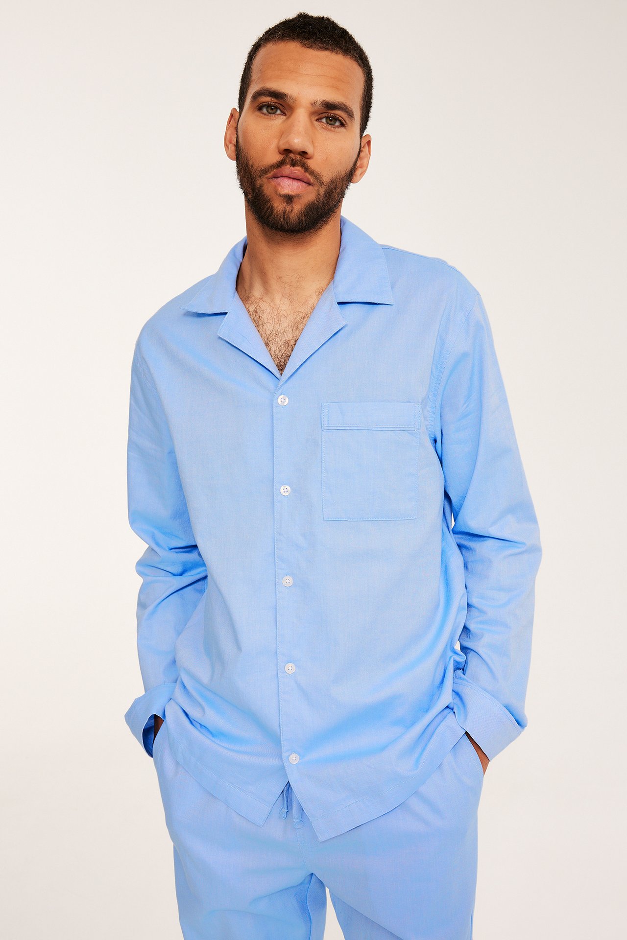 Framifrån, en man i en ljusblå pyjamas i Oxford-bomull med knäppt skjorta och bröstficka.