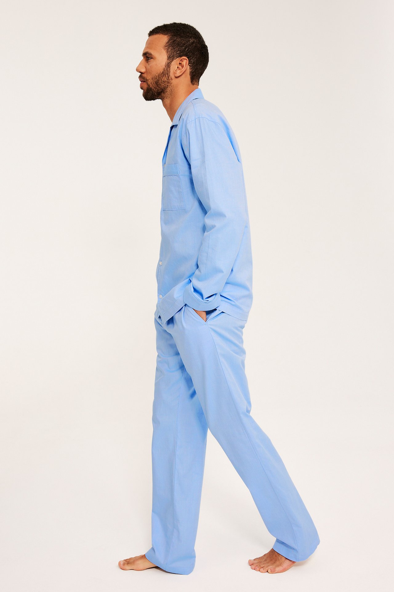 Sidovy av man i ljusblå Oxford bomullspyjamas med skjorta och byxor, händer i fickorna.